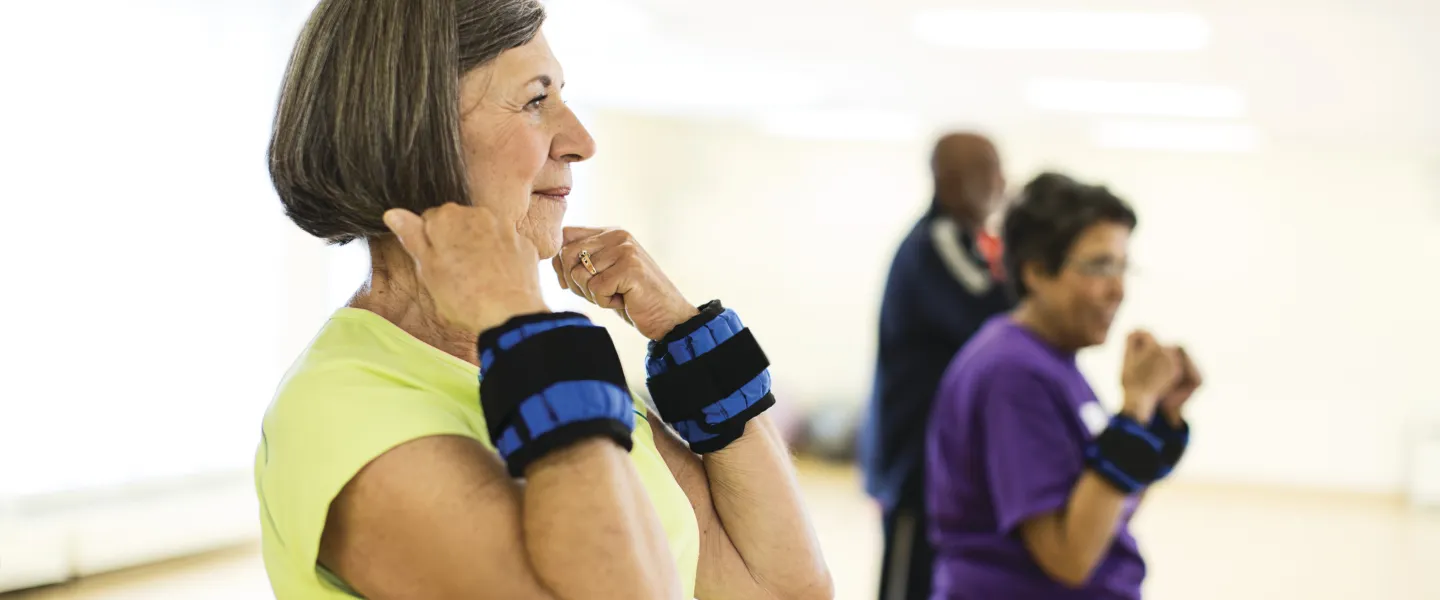 Senior women with wrist weights participate in a balance exercise class, other people are in the background, indoors.
