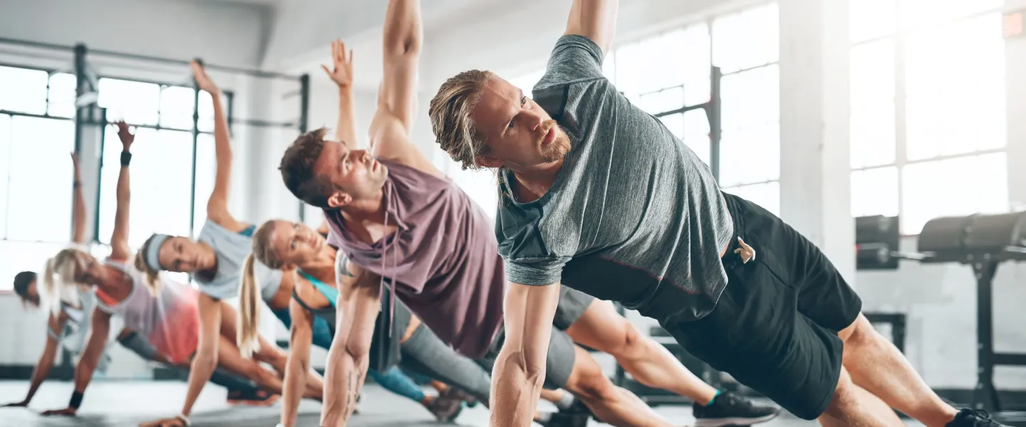 Group of people doing side planks with raised arms during fitness class in the gym. They are working on their core.
