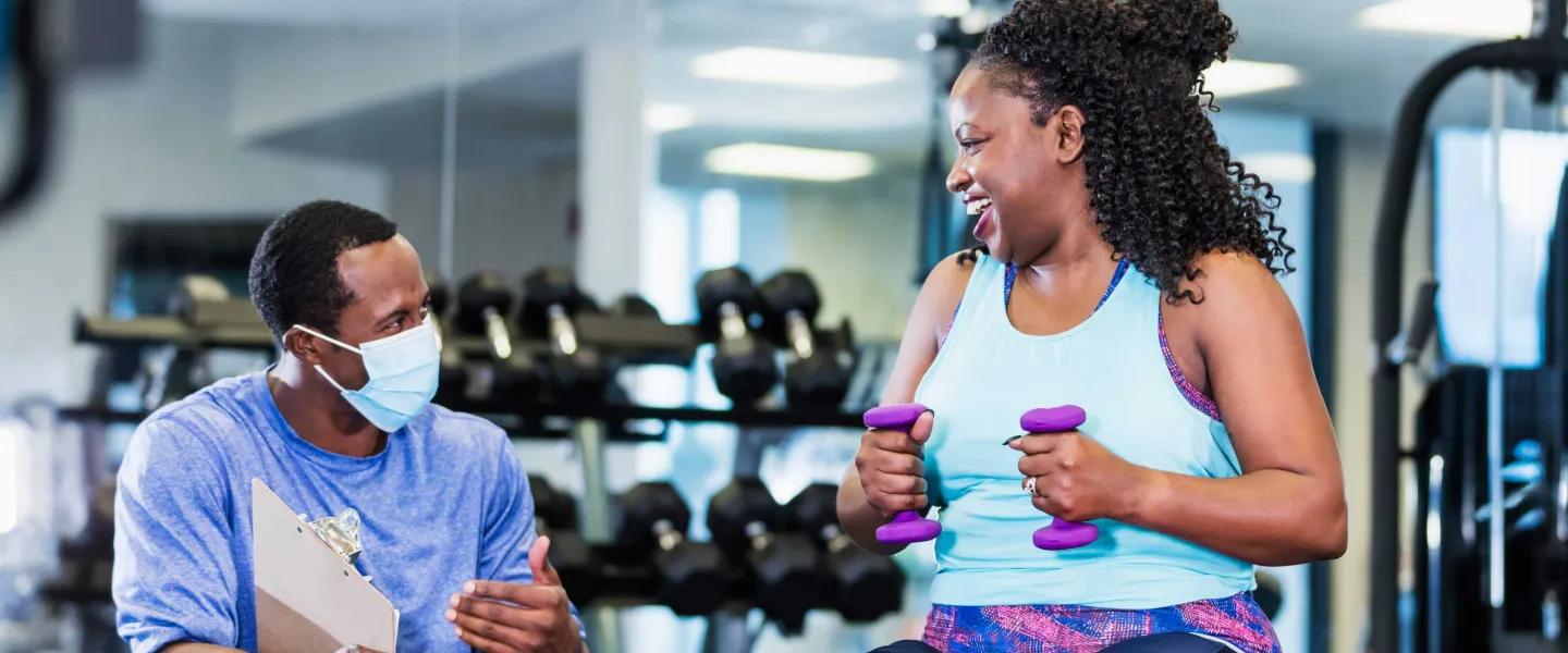 A trainer wearing a face mask coaches a woman exercising with weights, both laughing, in a fitness center. The trainer holds a clipboard.
