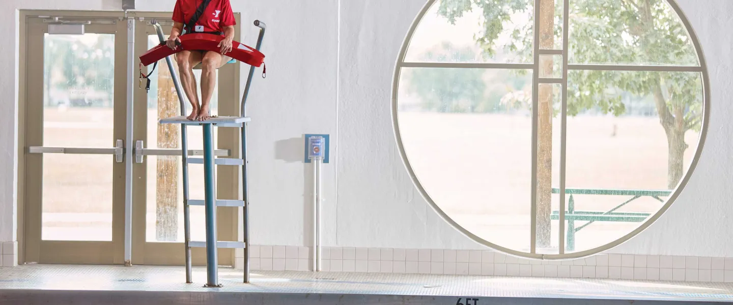 Lifeguard in red uniform sits atop a high chair, observing the pool; round window and door visible. The depth of the pool is marked with a 6ft sign.
