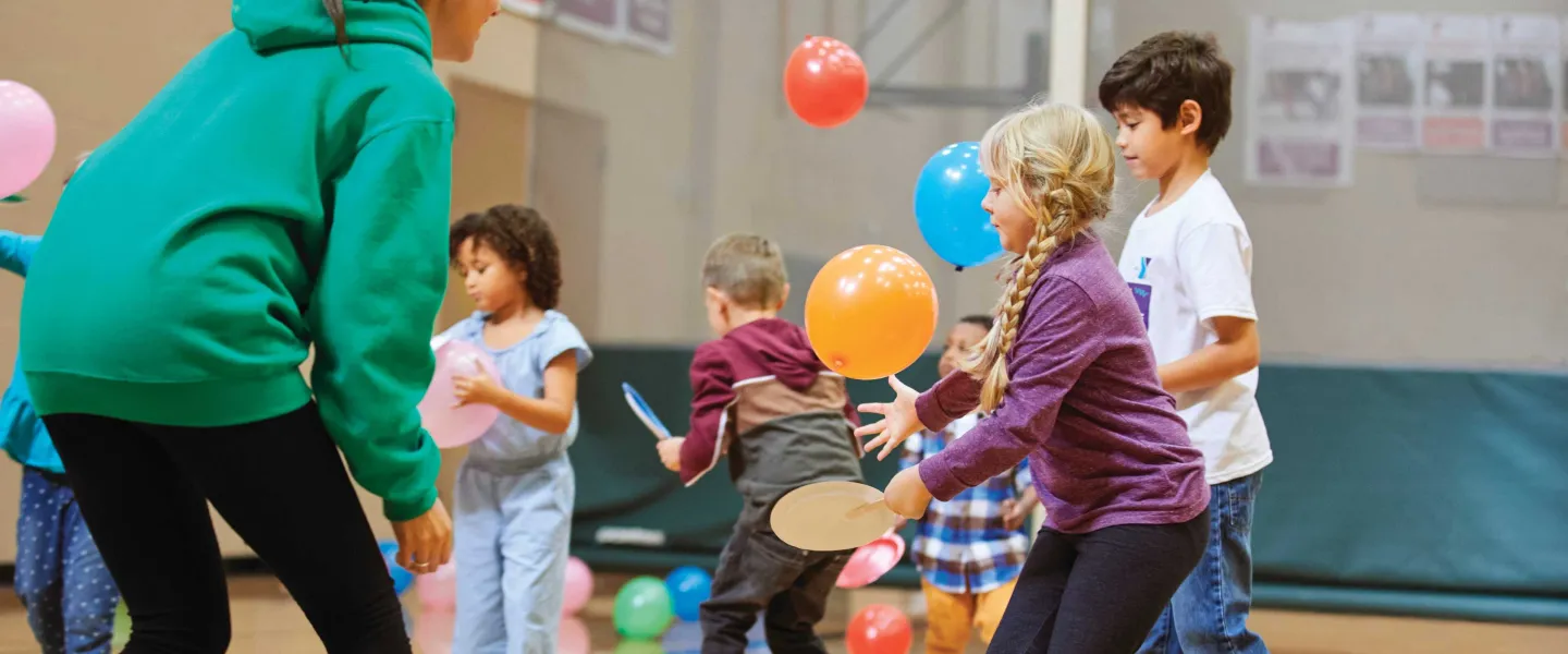 Children play with balloons and paddles during a recreation activity in a large gymnasium. A female counselor wearing a green hoodie supervises.
