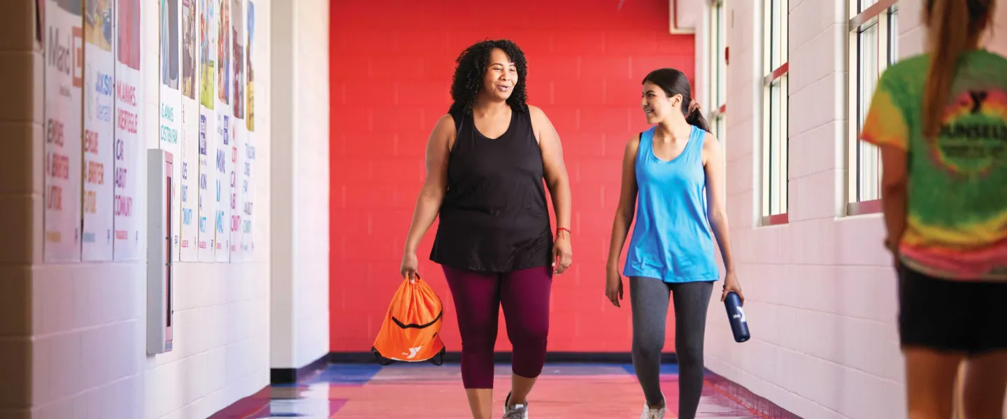 Two women are walking and talking in a brightly lit hallway with a third person walking away. One woman carries a bag, another a water bottle.