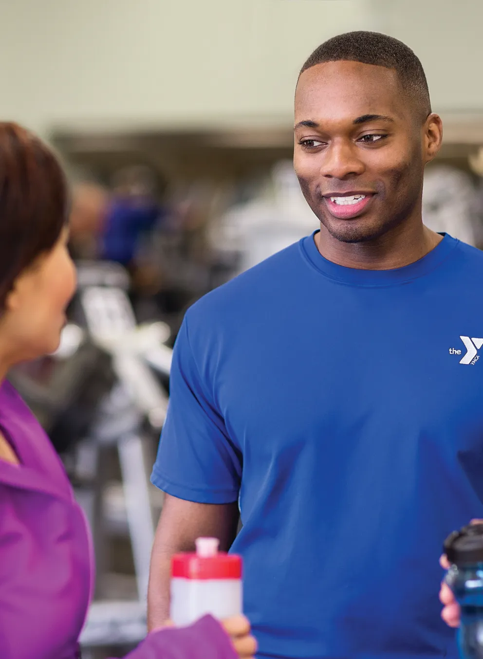 A staff member in a blue shirt with a logo chats with two members in a fitness center. They are holding water bottles and conversing.