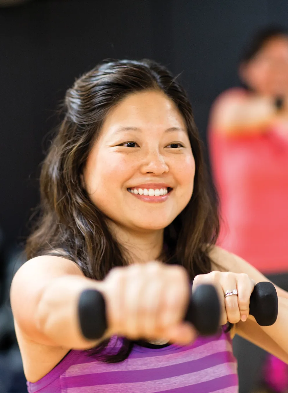 A smiling woman in a purple striped tank top holds dumbbells during a workout. Another person is blurred in the background.
