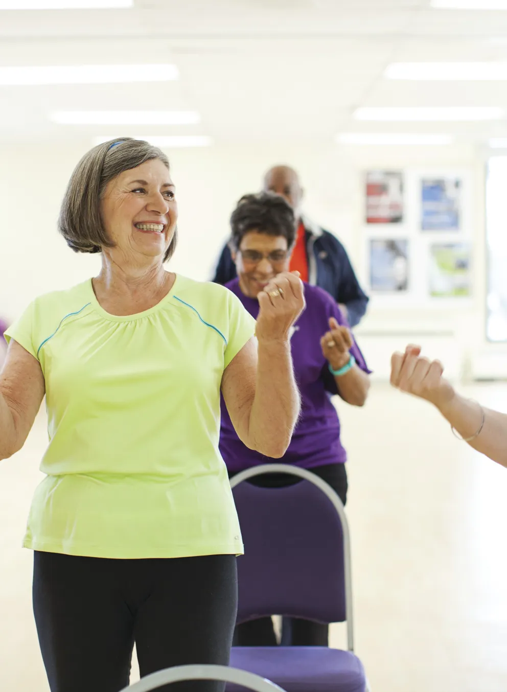 Active seniors participate in fitness class, smiling and raising their arms. People exercise in a light-filled room, seated on chairs.