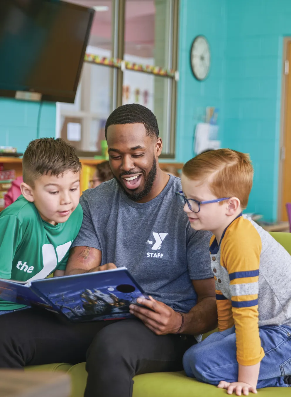 Adult staff member reads a book to two young children, seated on a green couch. The atmosphere is in a youth center. A sign indicates daily activities.