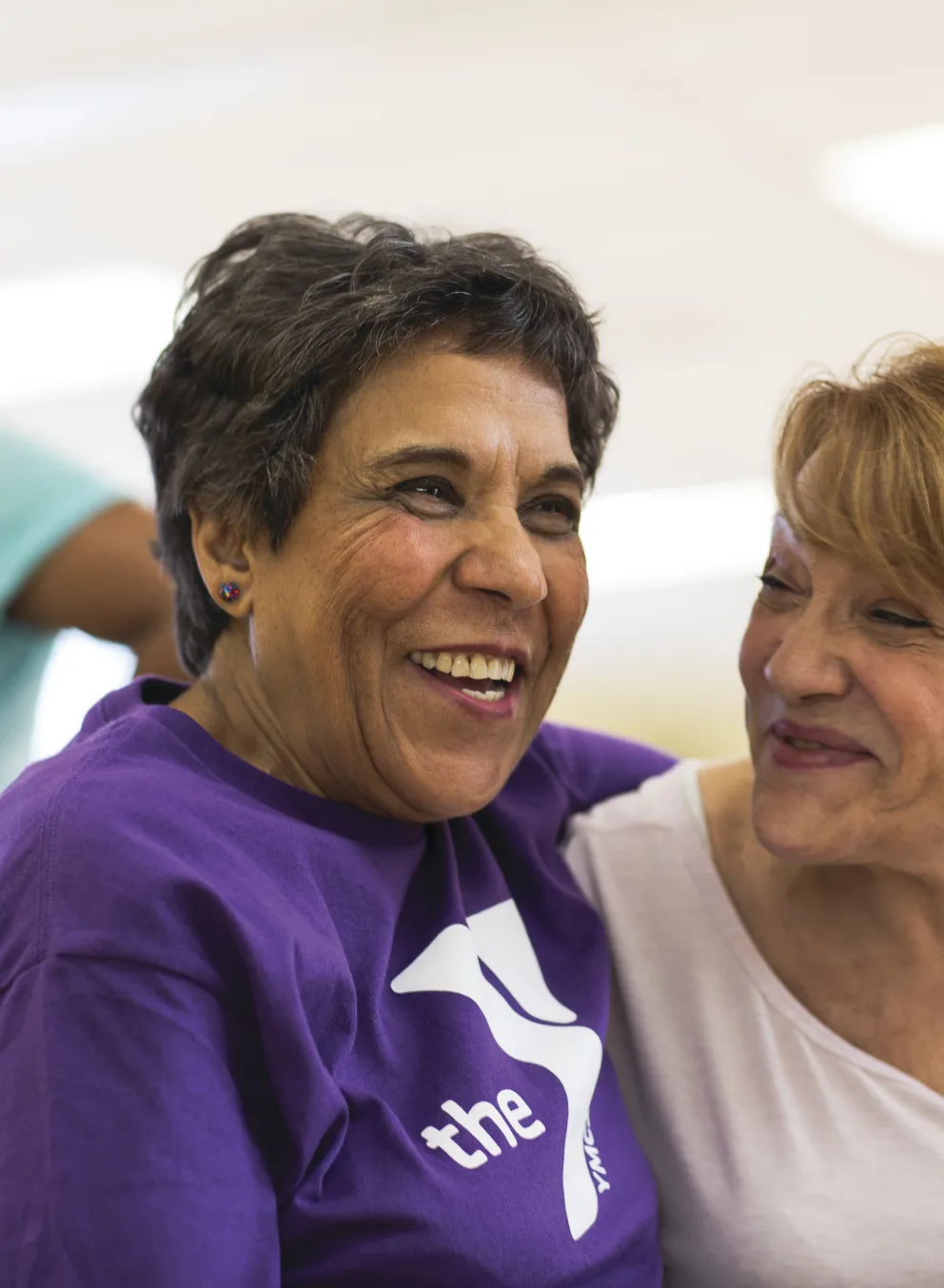 Two smiling women with arms around each other, one in purple shirt with logo, another woman in the background.