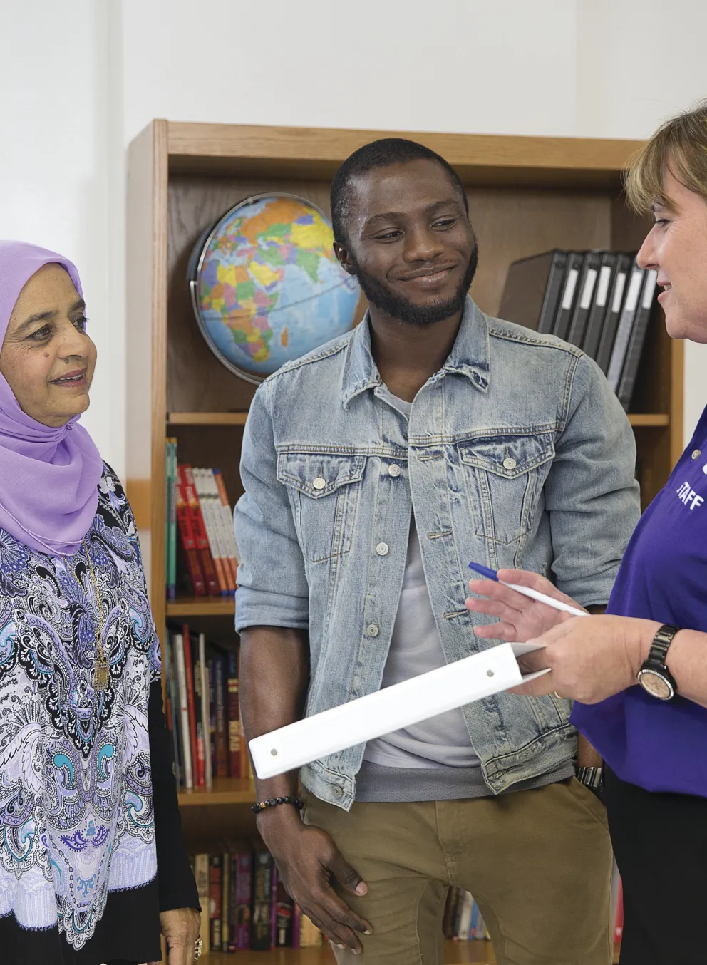 Three adults in a community setting are engaged in a discussion, with a woman holding a whiteboard. A bookshelf and world globe are visible.