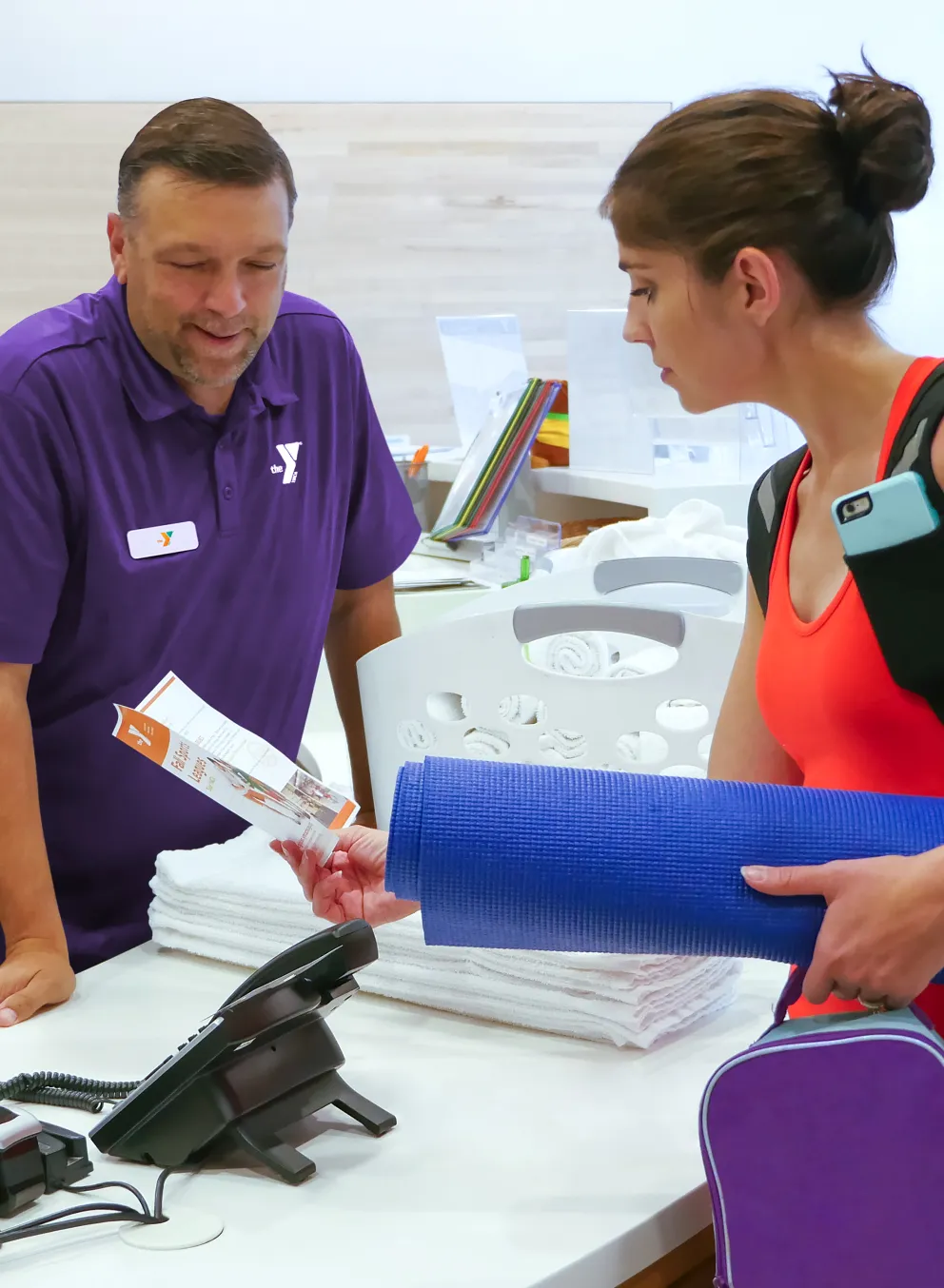 A staff member hands paperwork to a woman with a yoga mat and backpack at a community center reception desk; other people wait nearby.