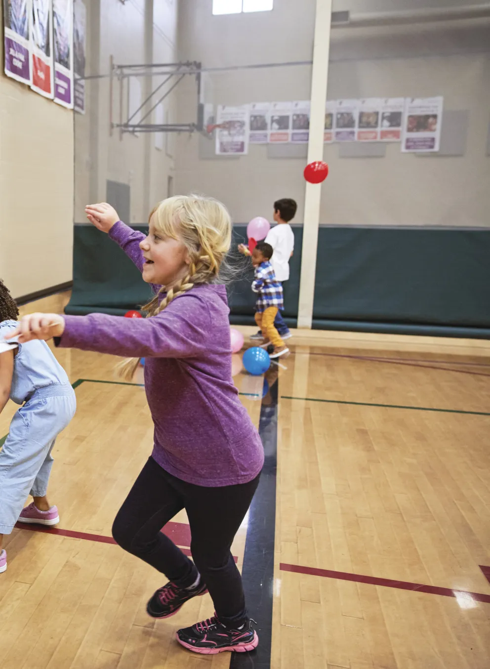 Children playing games in a gymnasium, with an instructor holding a paper strip. Kids enjoy active recreation, smiling and laughing. Colorful balloons scattered on the wooden floor.
