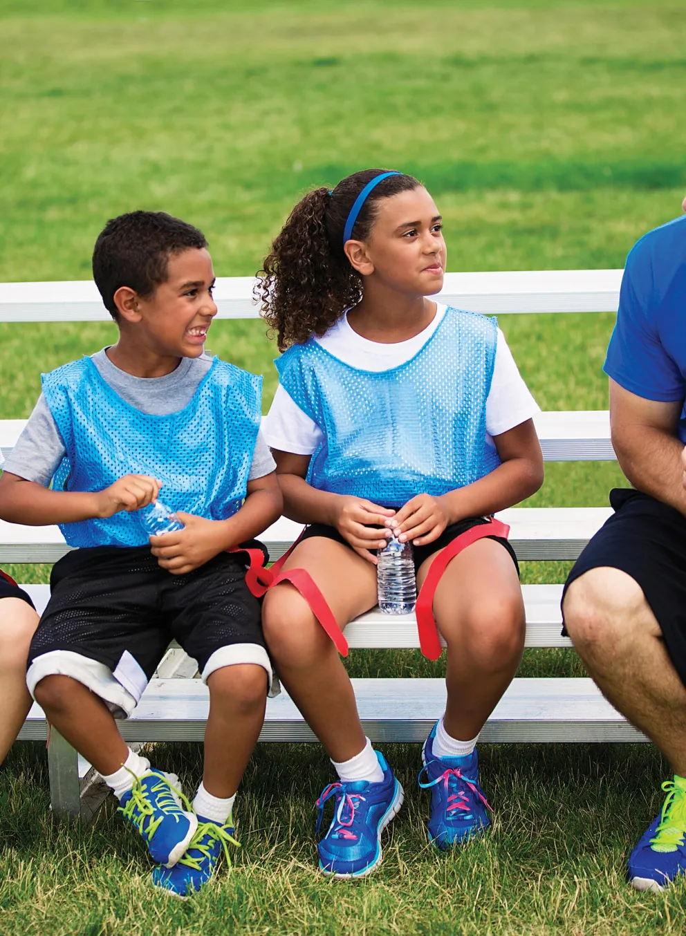 Four kids and a coach sit on a bench on a grass field. The coach holds a football. Kids wear vests and listen.