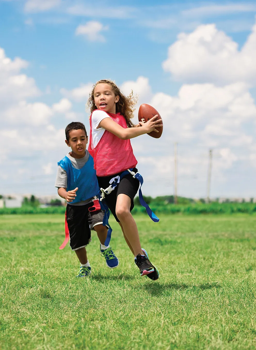 Children playing flag football on a grassy field, girl throwing a football, boy running beside her, other kids watching.