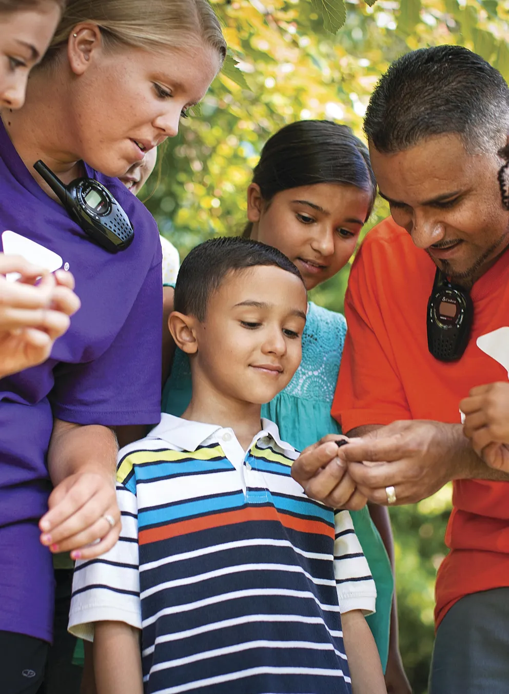 Adults and children gather around, examining something small in the open air. They are looking at an object together. The shirts have a logo.