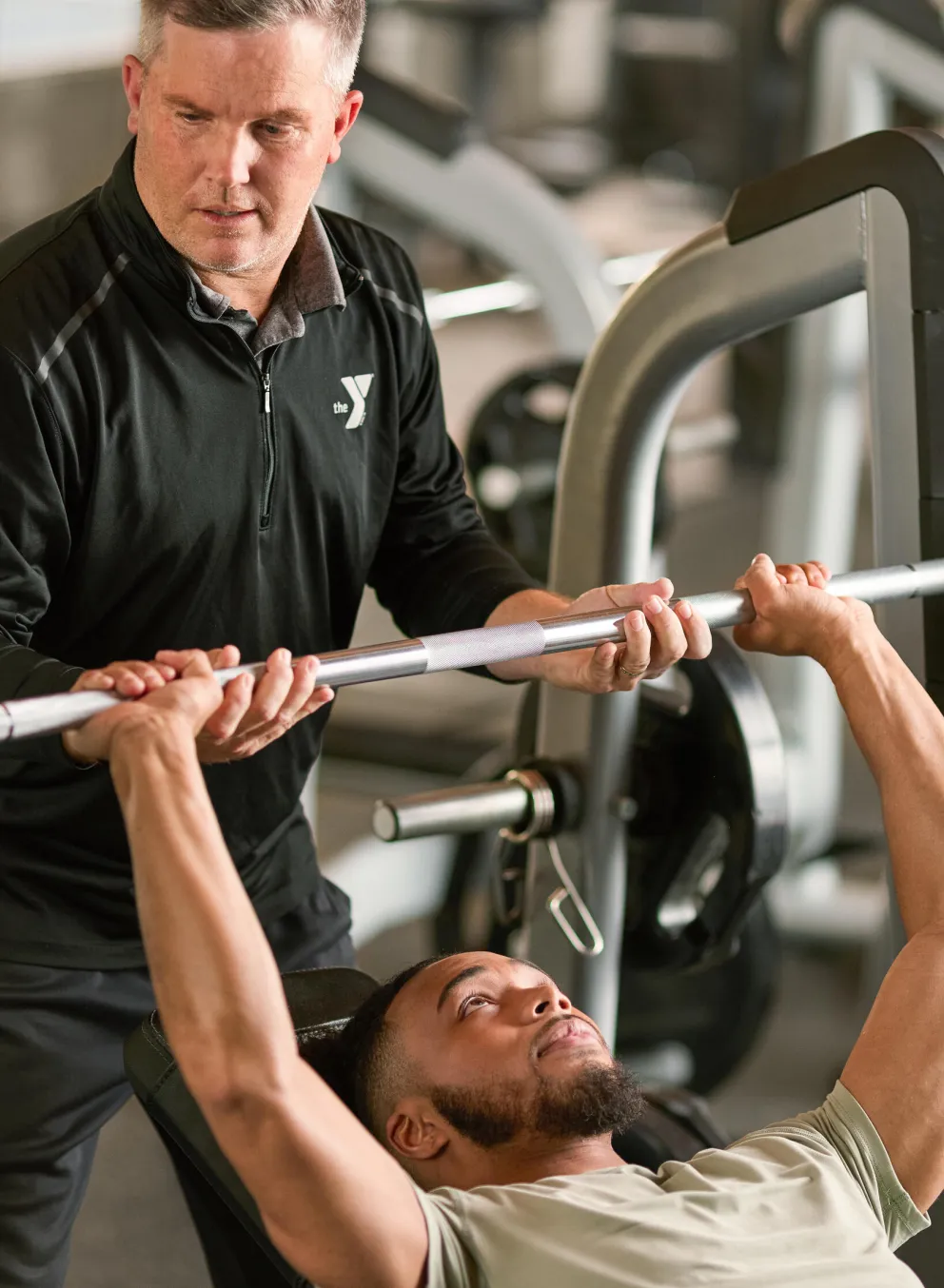 A man receives guidance from a fitness instructor while bench pressing a barbell in a gym.