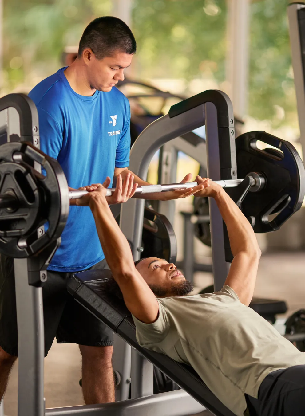 A man bench presses a weighted barbell, assisted by a trainer in a bright blue shirt.  Other fitness equipment is visible in the background.