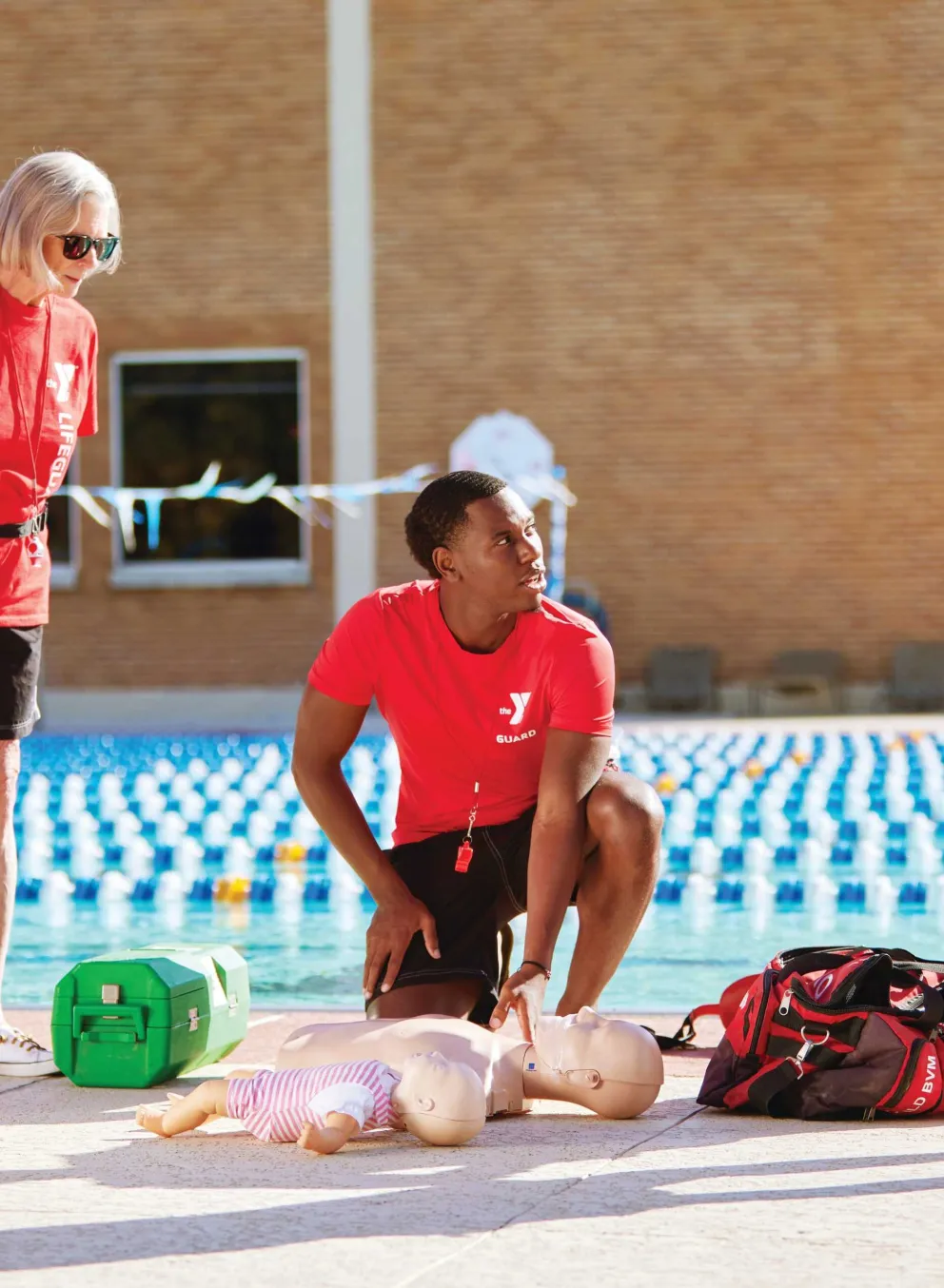 Lifeguards in red shirts practice CPR on a training dummy by the pool with others watching. A green first aid kit and a red bag lay nearby.