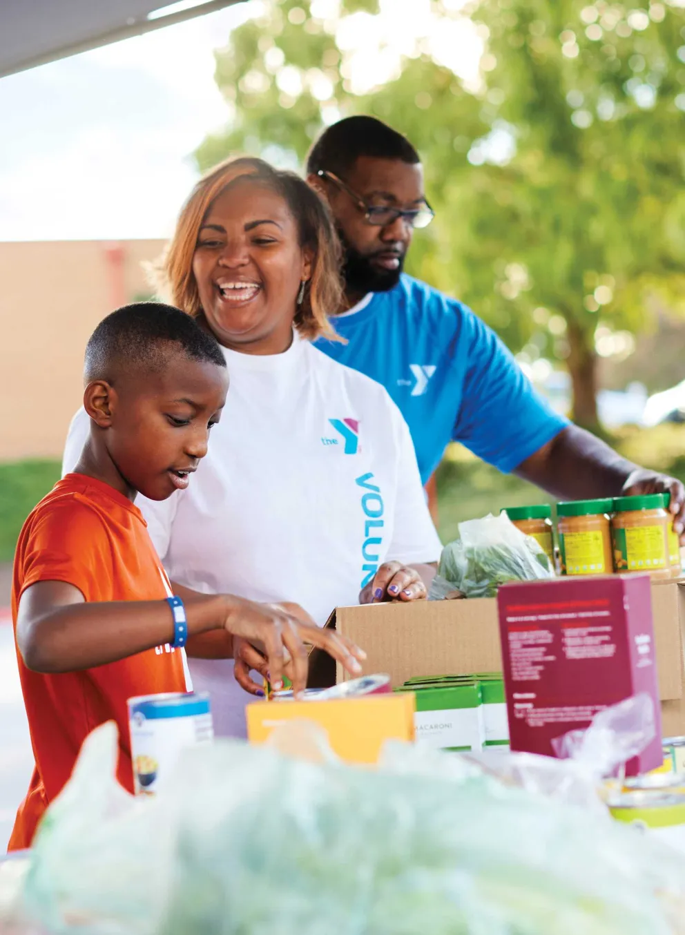 Volunteers and a young boy packing food in boxes at an outdoor community event; they are sorting items and smiling.