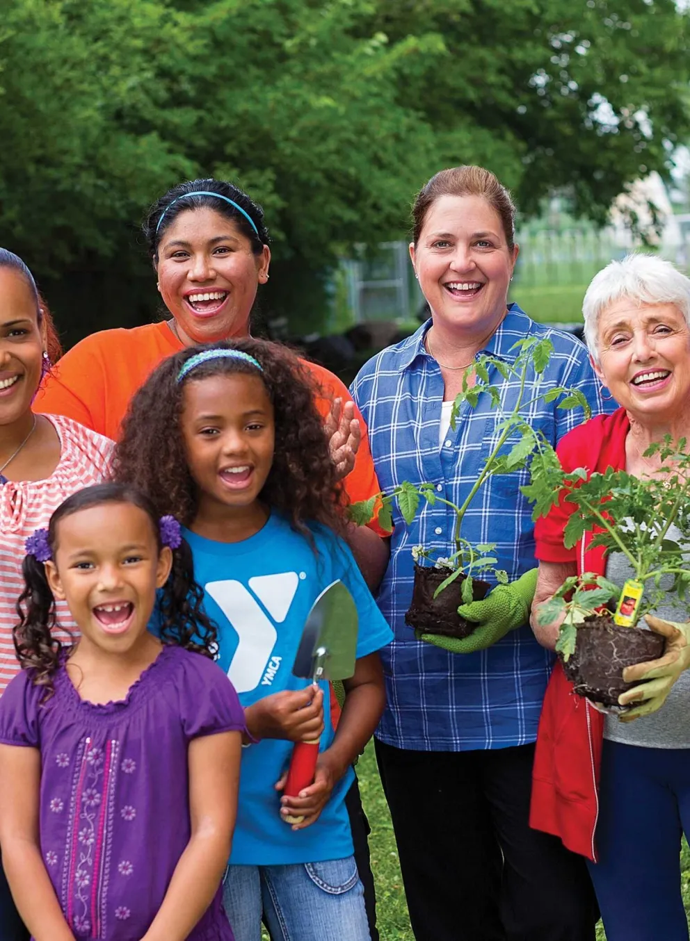 Group of diverse people smile, posing for a photo together in the garden. Kids and adults hold plants, gardening tools; a community activity.
