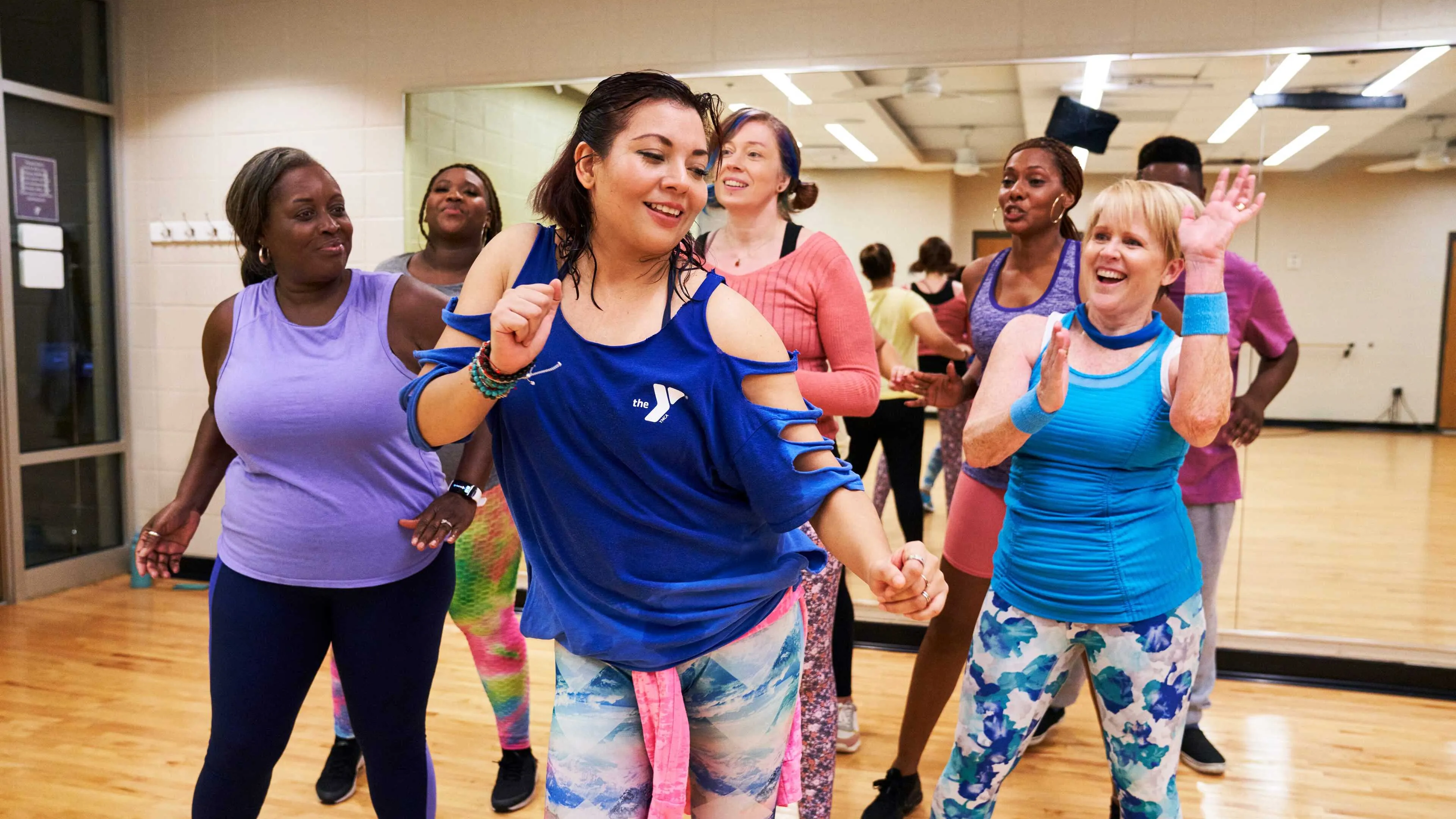 Group of people are in a studio, wearing activewear, dancing during a fitness class, and appear joyful. Brightly colored outfits are worn by participants.