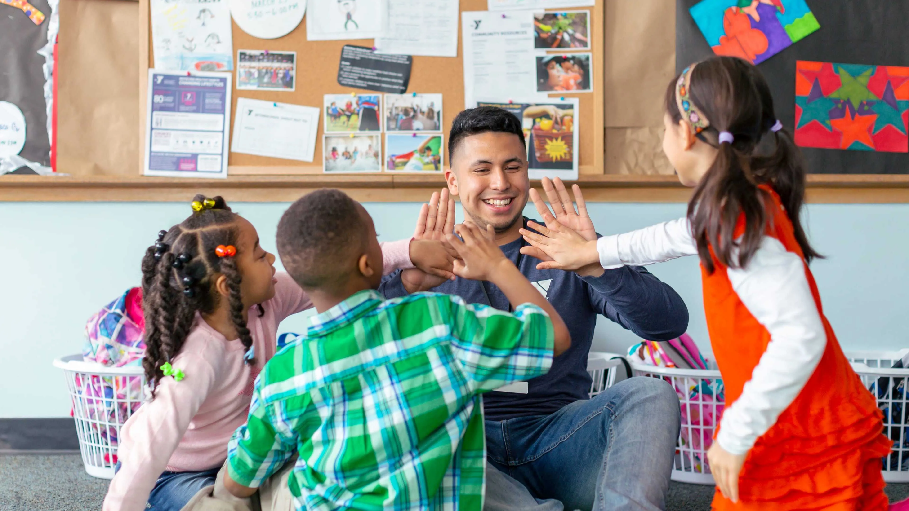 A counselor and four children in a circle sit on the floor, all giving high-fives in a playful classroom scene.