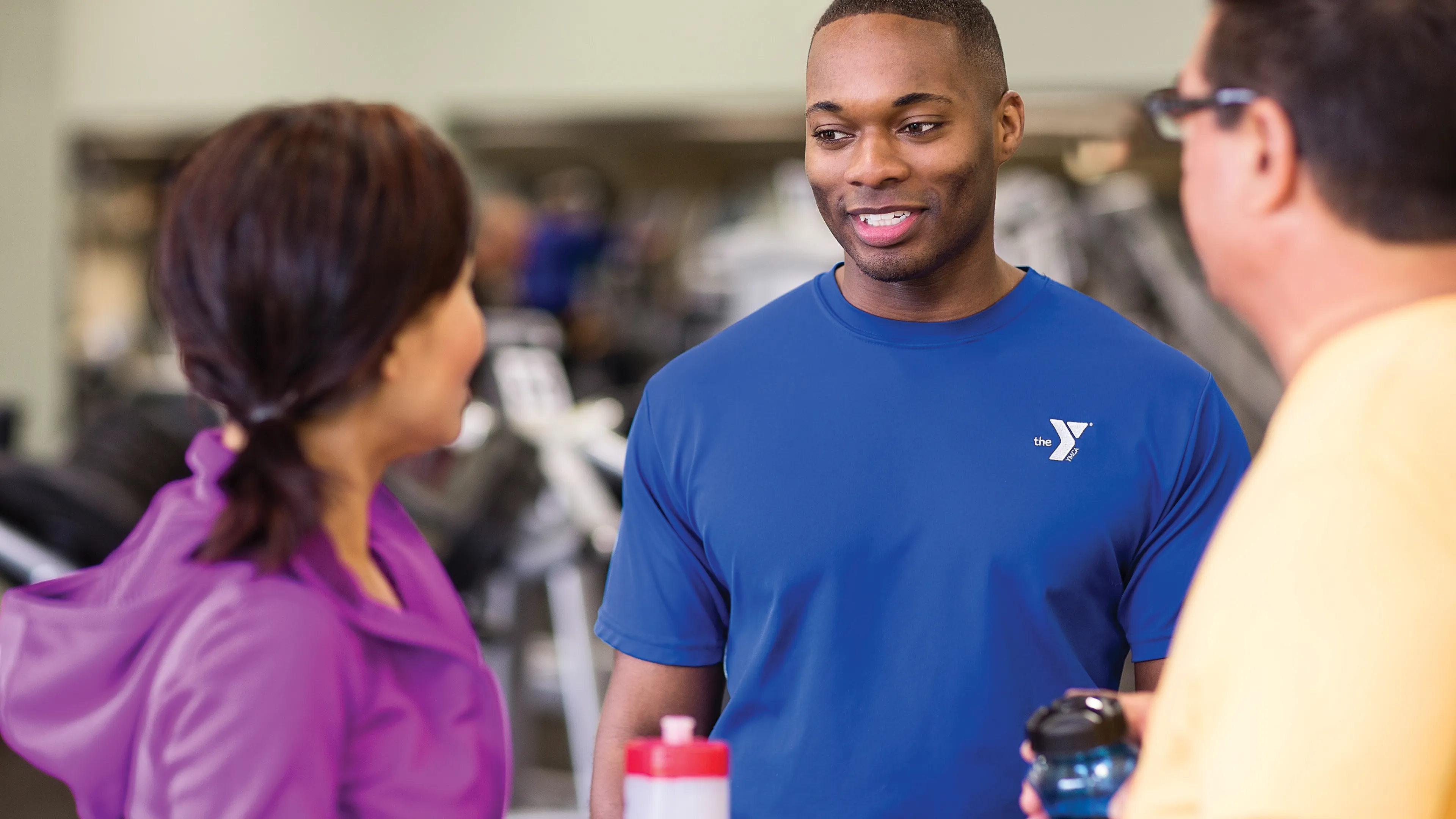 A staff member in a blue shirt with a logo chats with two members in a fitness center. They are holding water bottles and conversing.