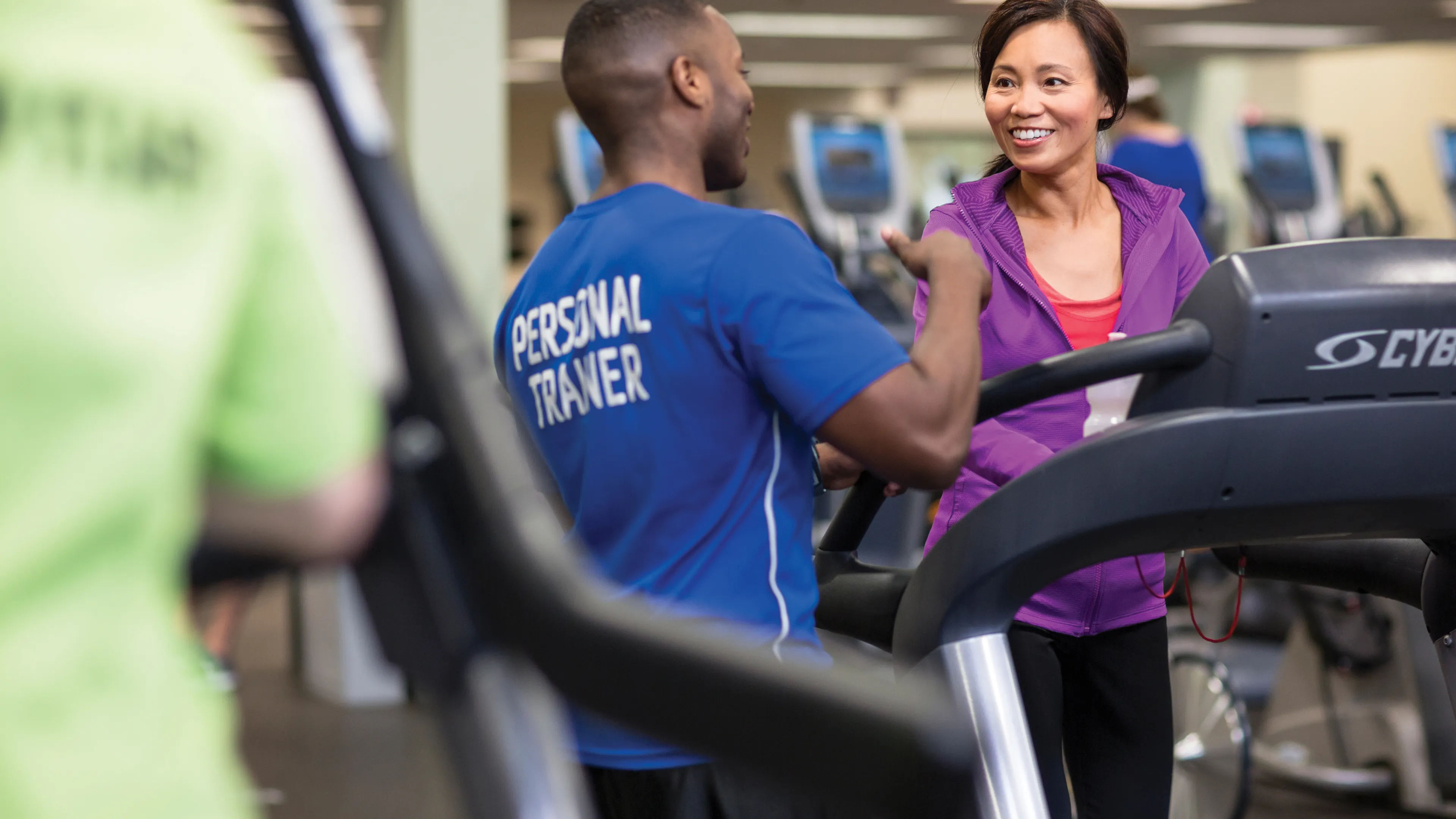 Personal trainer in blue shirt talking with a woman on treadmill; wellness center workout is ongoing. Another person is running on a machine.