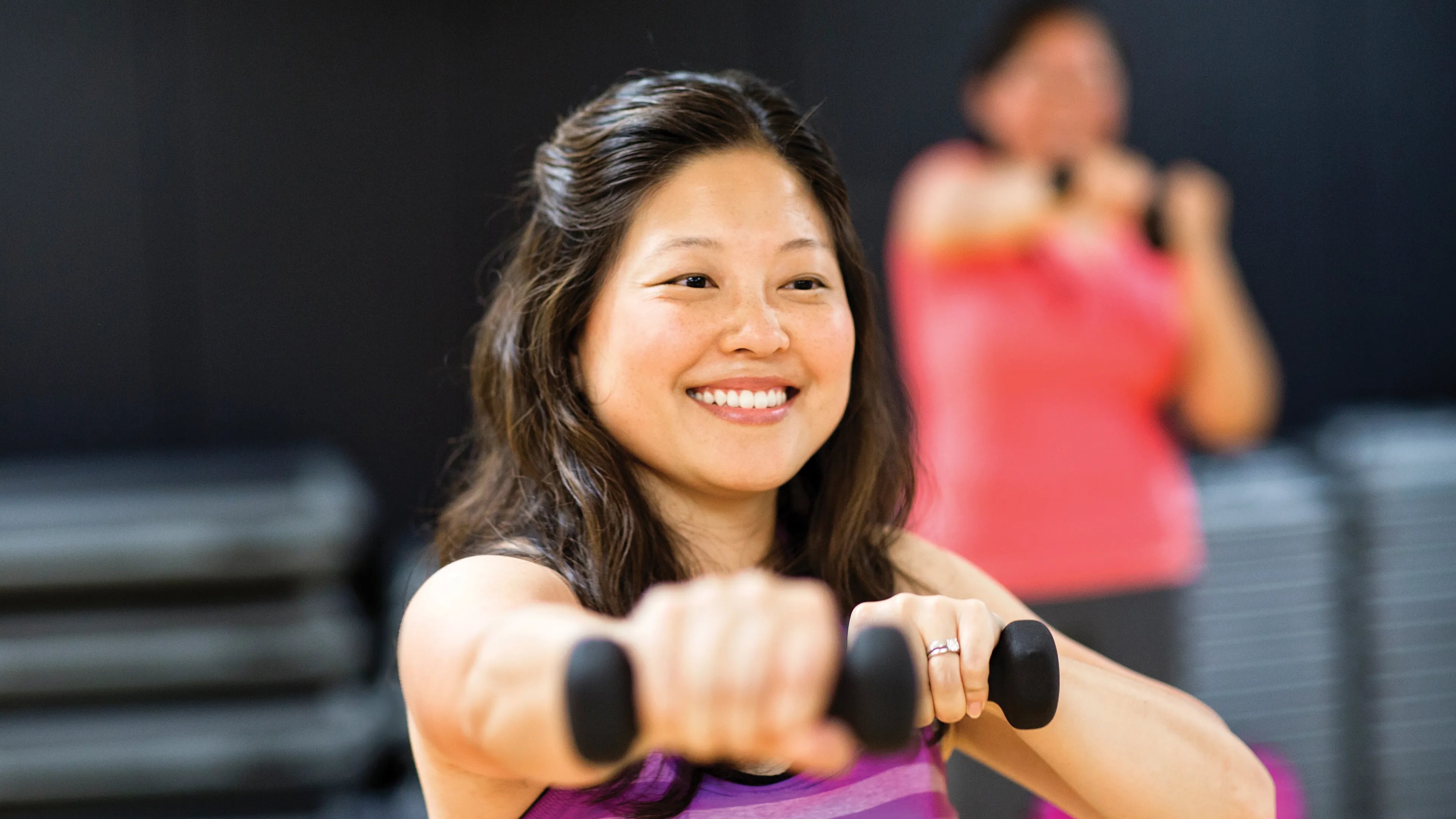 A smiling woman in a purple striped tank top holds dumbbells during a workout. Another person is blurred in the background.