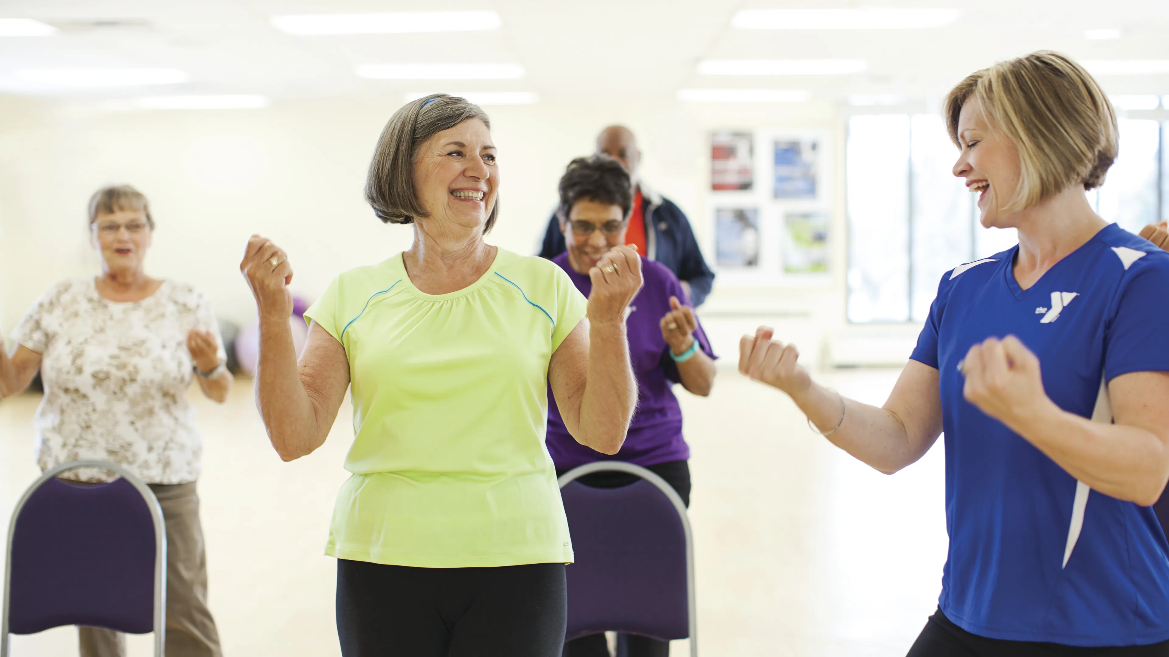 Active seniors participate in fitness class, smiling and raising their arms. People exercise in a light-filled room, seated on chairs.