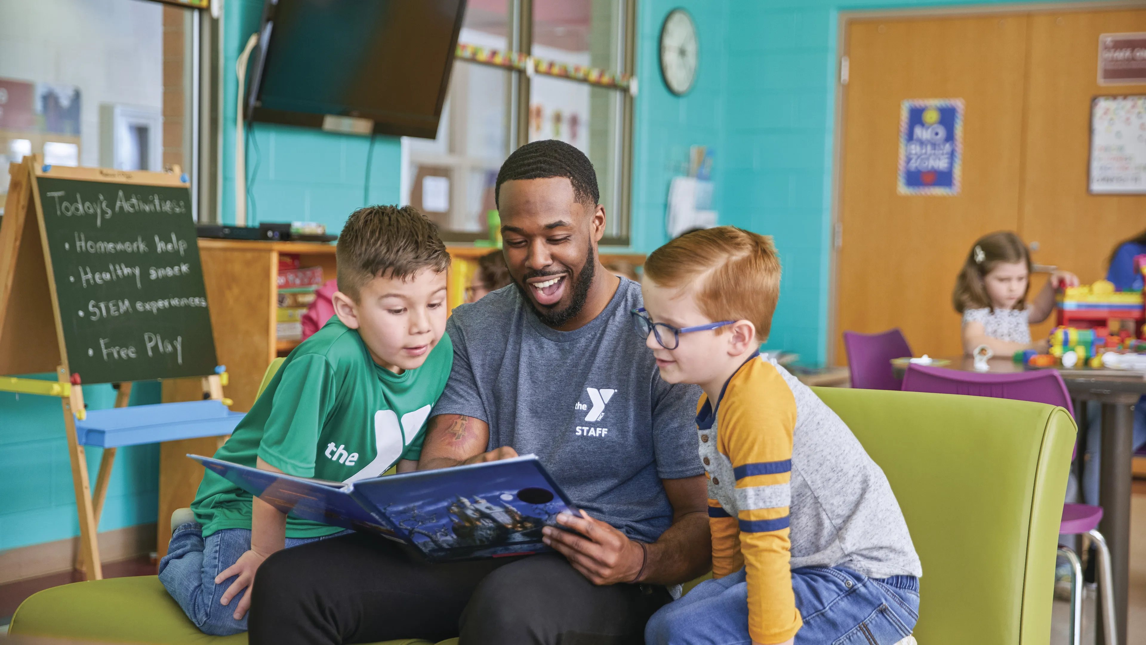 Adult staff member reads a book to two young children, seated on a green couch. The atmosphere is in a youth center. A sign indicates daily activities.