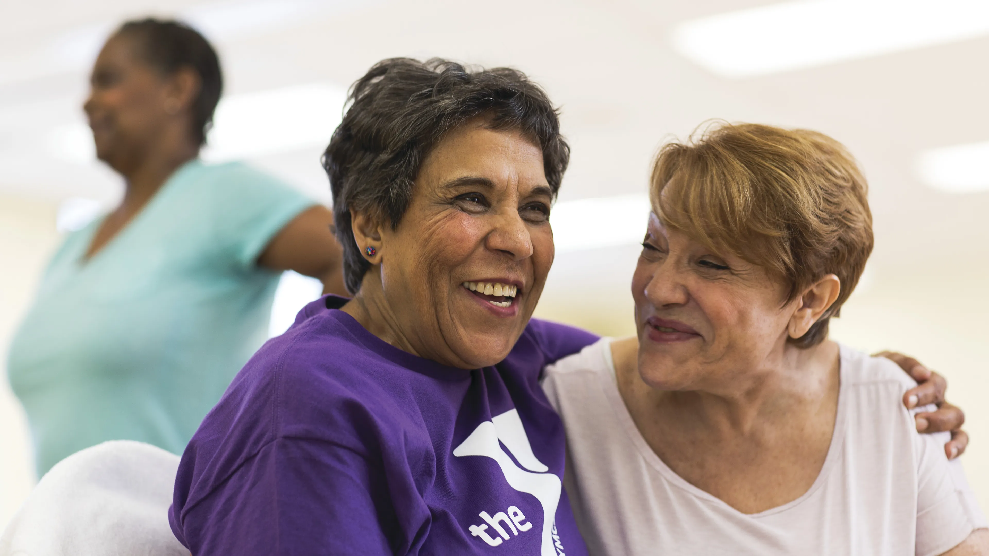 Two smiling women with arms around each other, one in purple shirt with logo, another woman in the background.