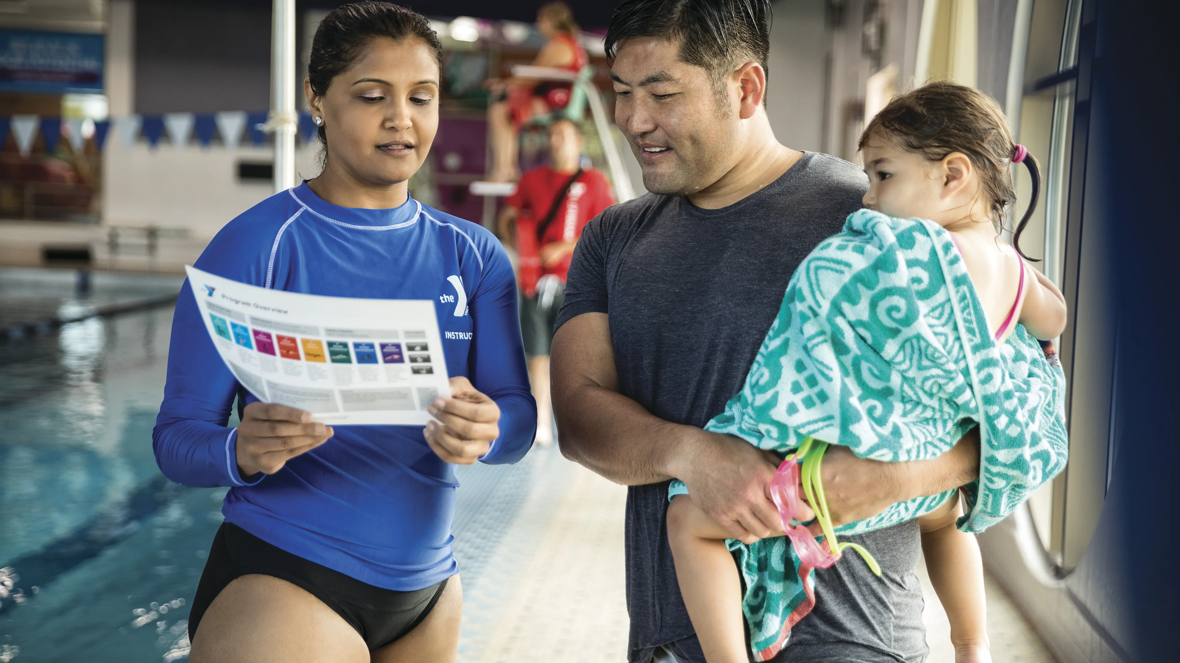 Instructor in blue shirt holding a program paper discussing with a father, holding a toddler wrapped in a towel, by the indoor pool.