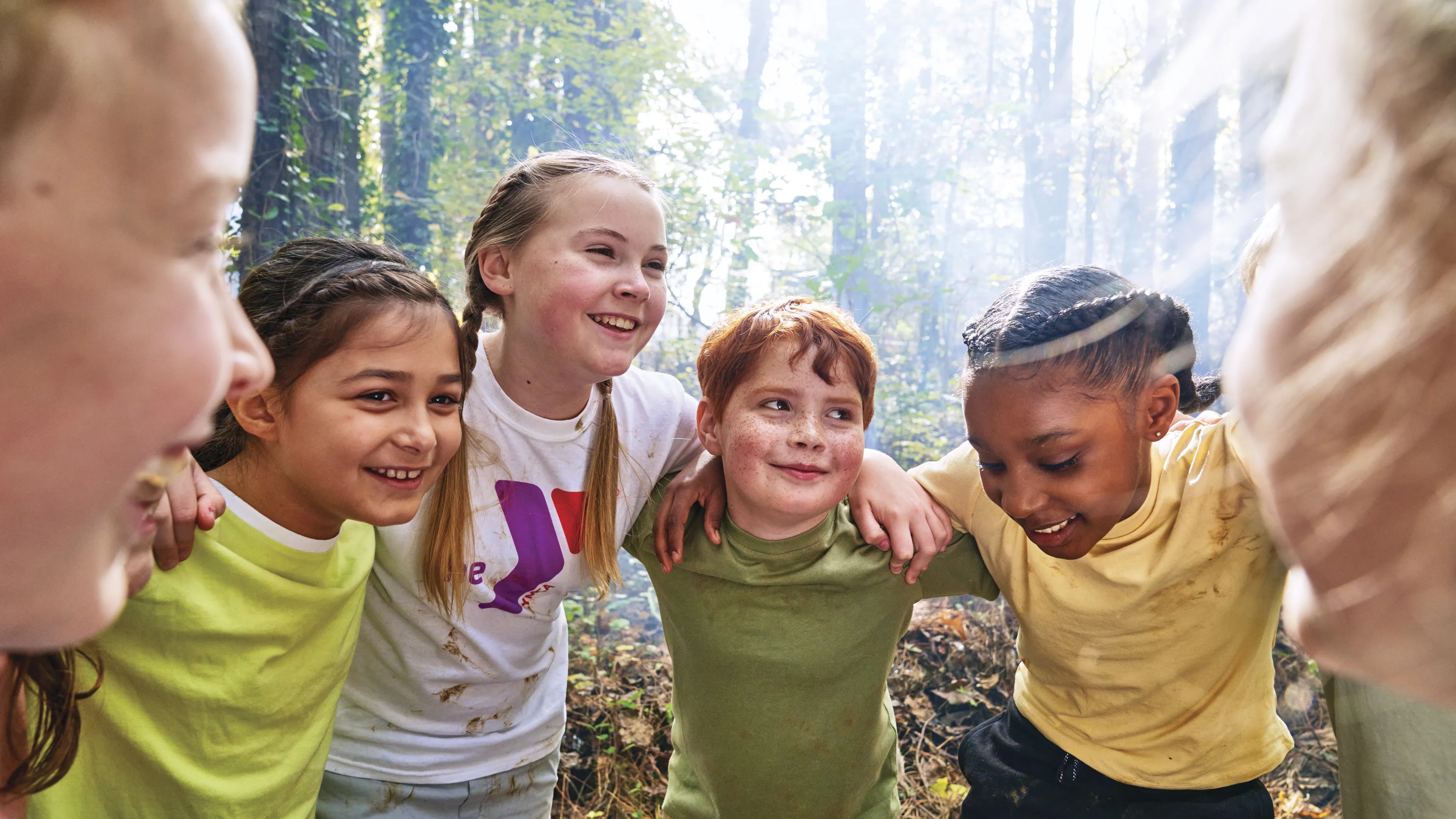 Group of kids huddled together, smiling, with arms around each other in a forest setting, enjoying camp activities.