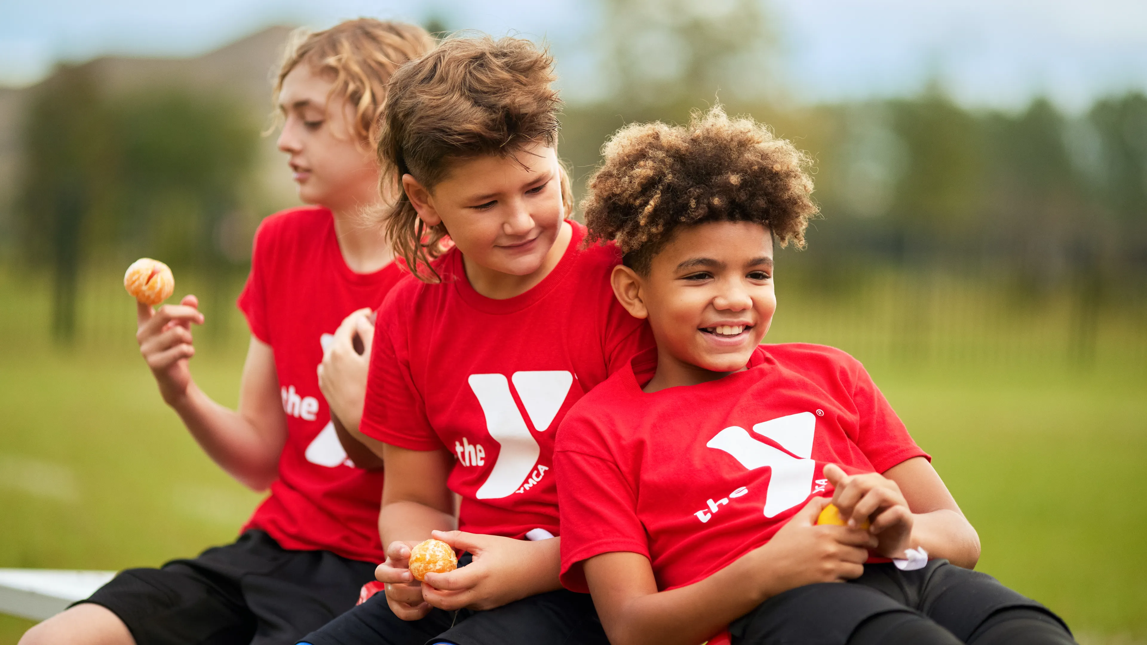 Three young children in red shirts with white logos and black shorts smile, holding small balls while sitting outside on a bench.