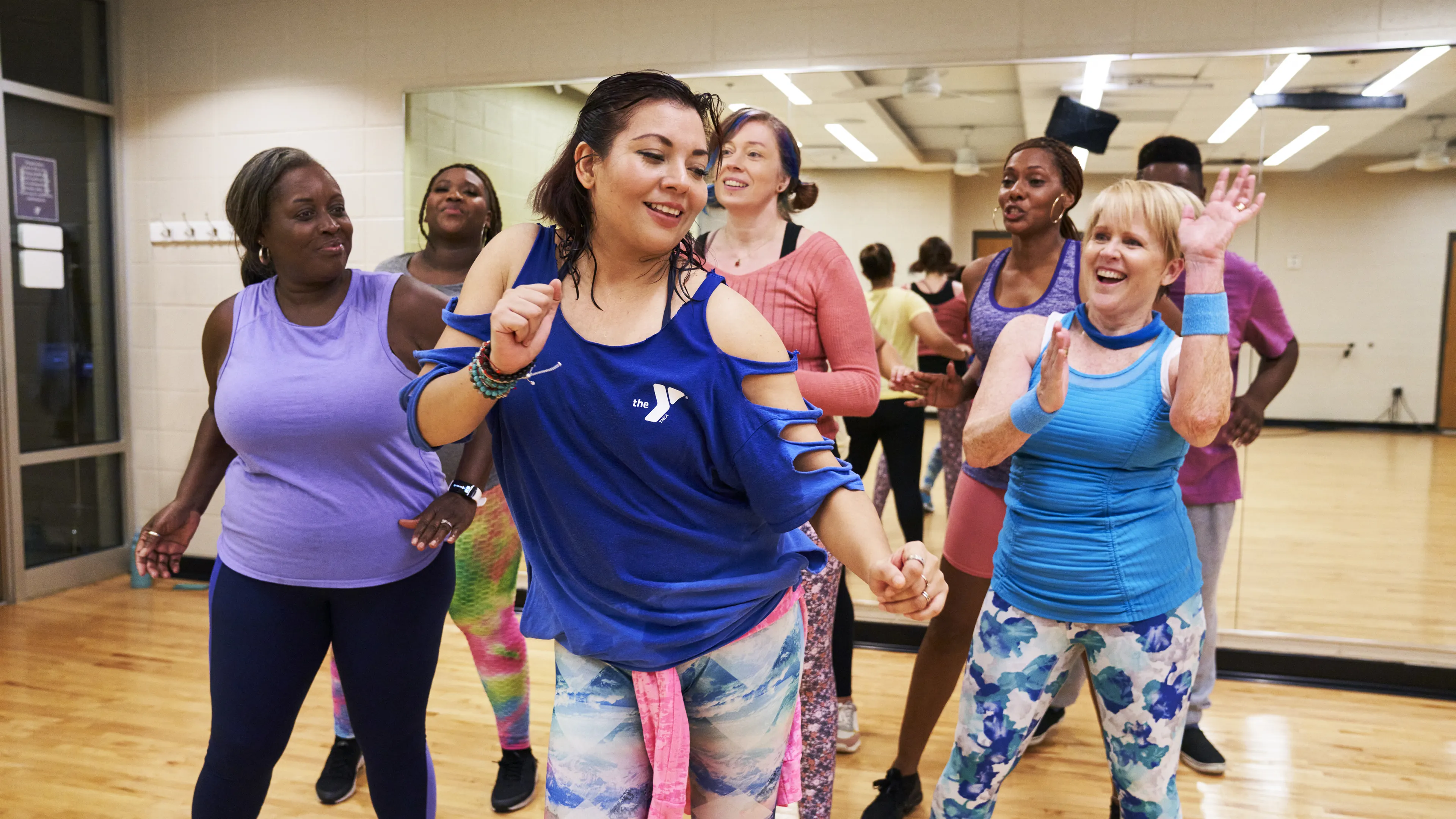 A group of diverse people in athletic attire dance joyfully in a fitness class, holding hands, and smiling broadly.