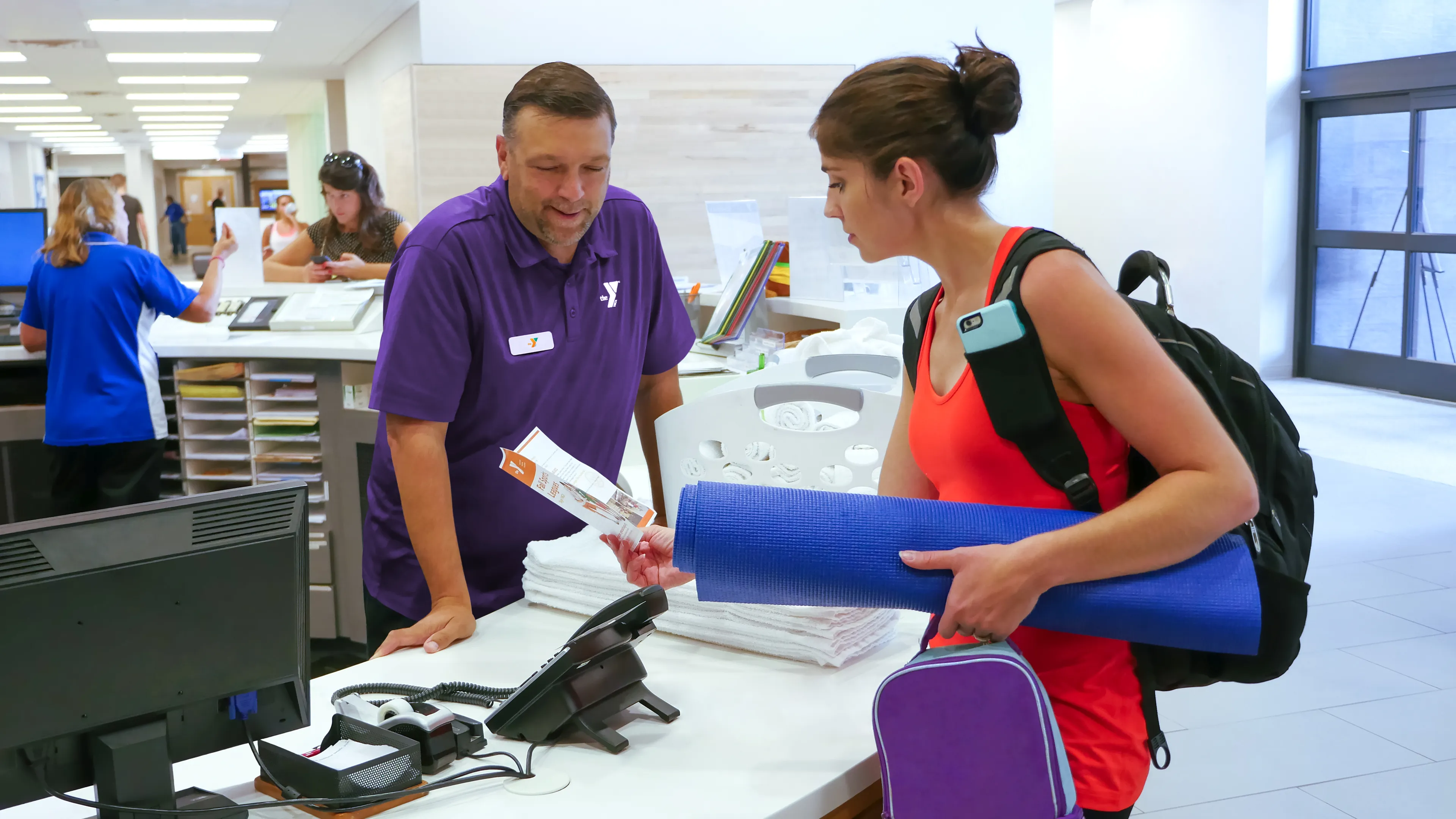 A staff member hands paperwork to a woman with a yoga mat and backpack at a community center reception desk; other people wait nearby.