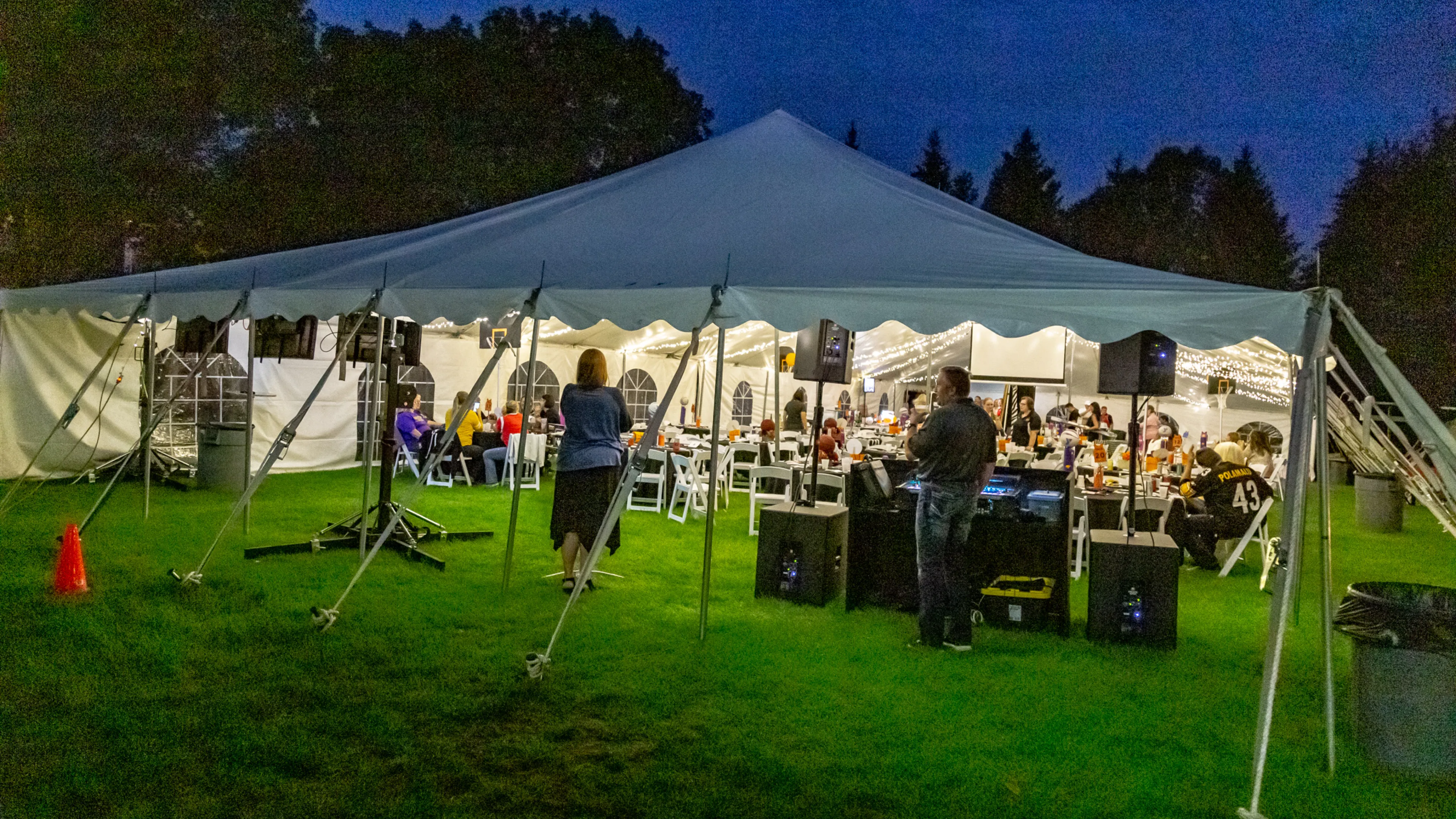 Large white tent set on green grass, tables and people inside, a DJ playing music for a community event at night.