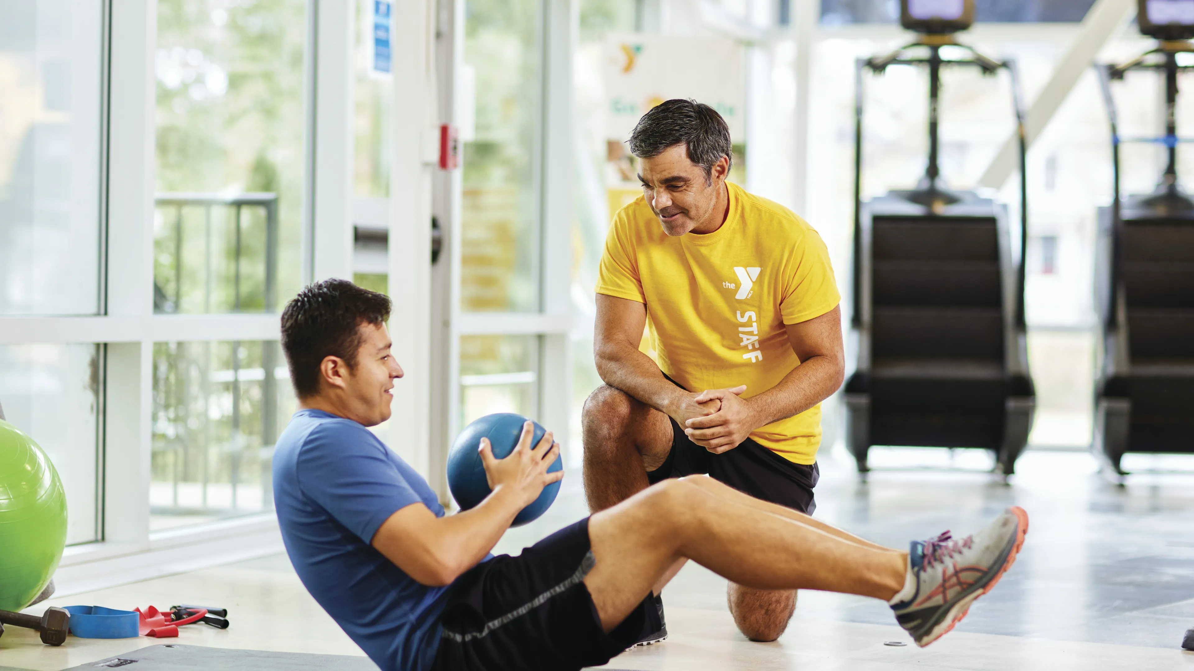 Man doing core workout with a medicine ball while a trainer kneels nearby, watching the exercise.