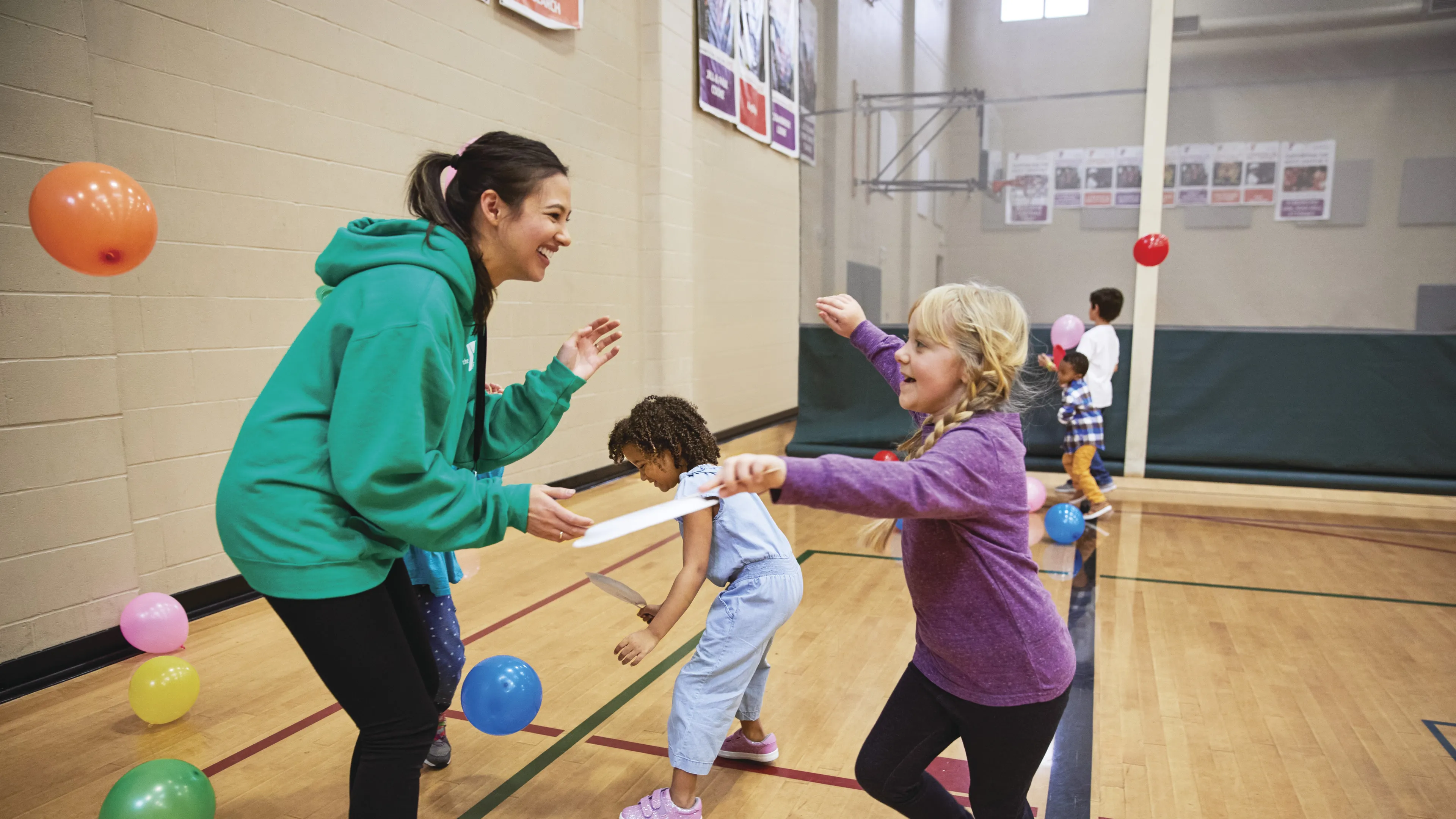 Children playing games in a gymnasium, with an instructor holding a paper strip. Kids enjoy active recreation, smiling and laughing. Colorful balloons scattered on the wooden floor.