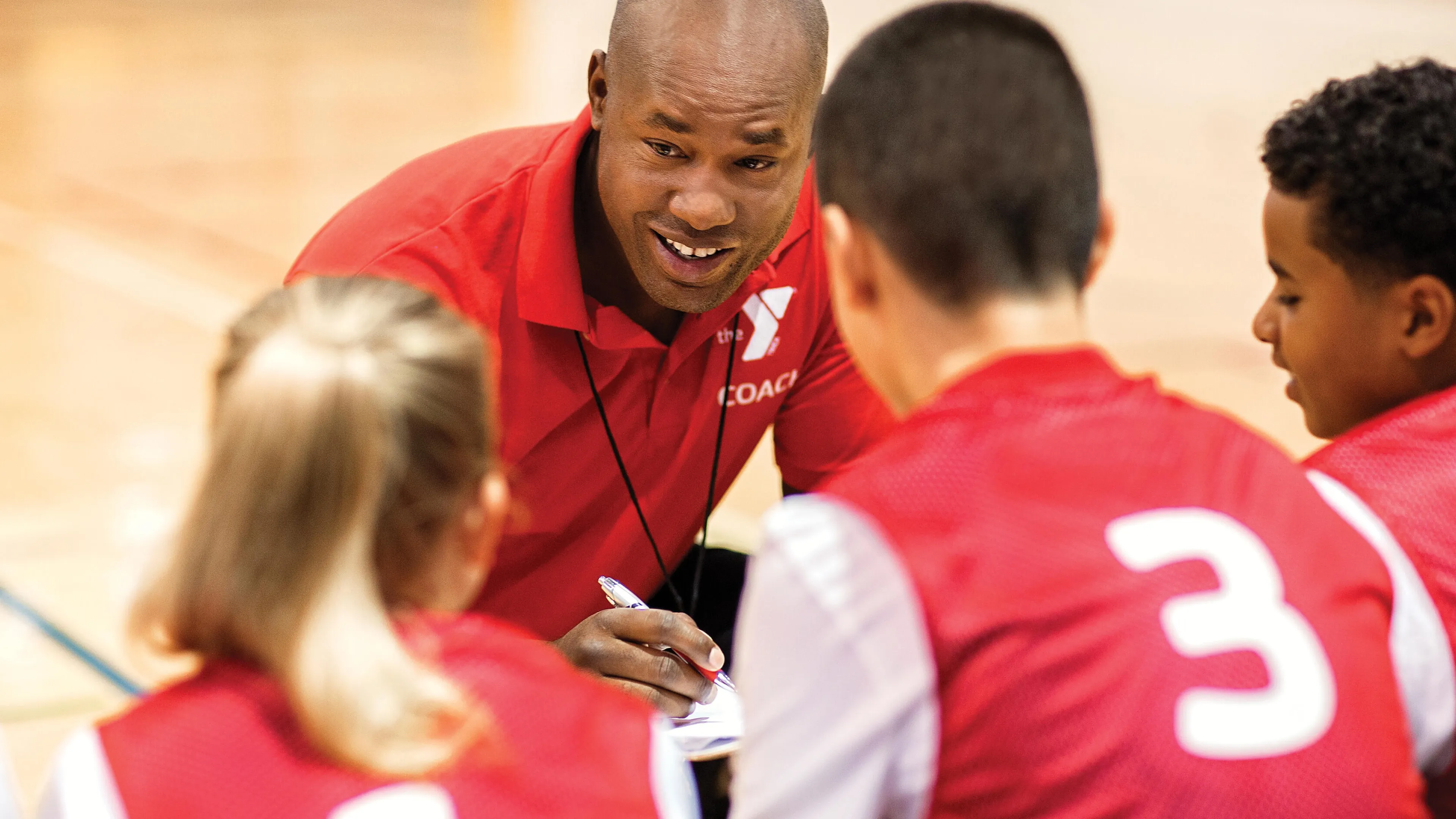 A coach in a red shirt with "COACH" talks to kids in red team shirts numbered one and three on a basketball court.