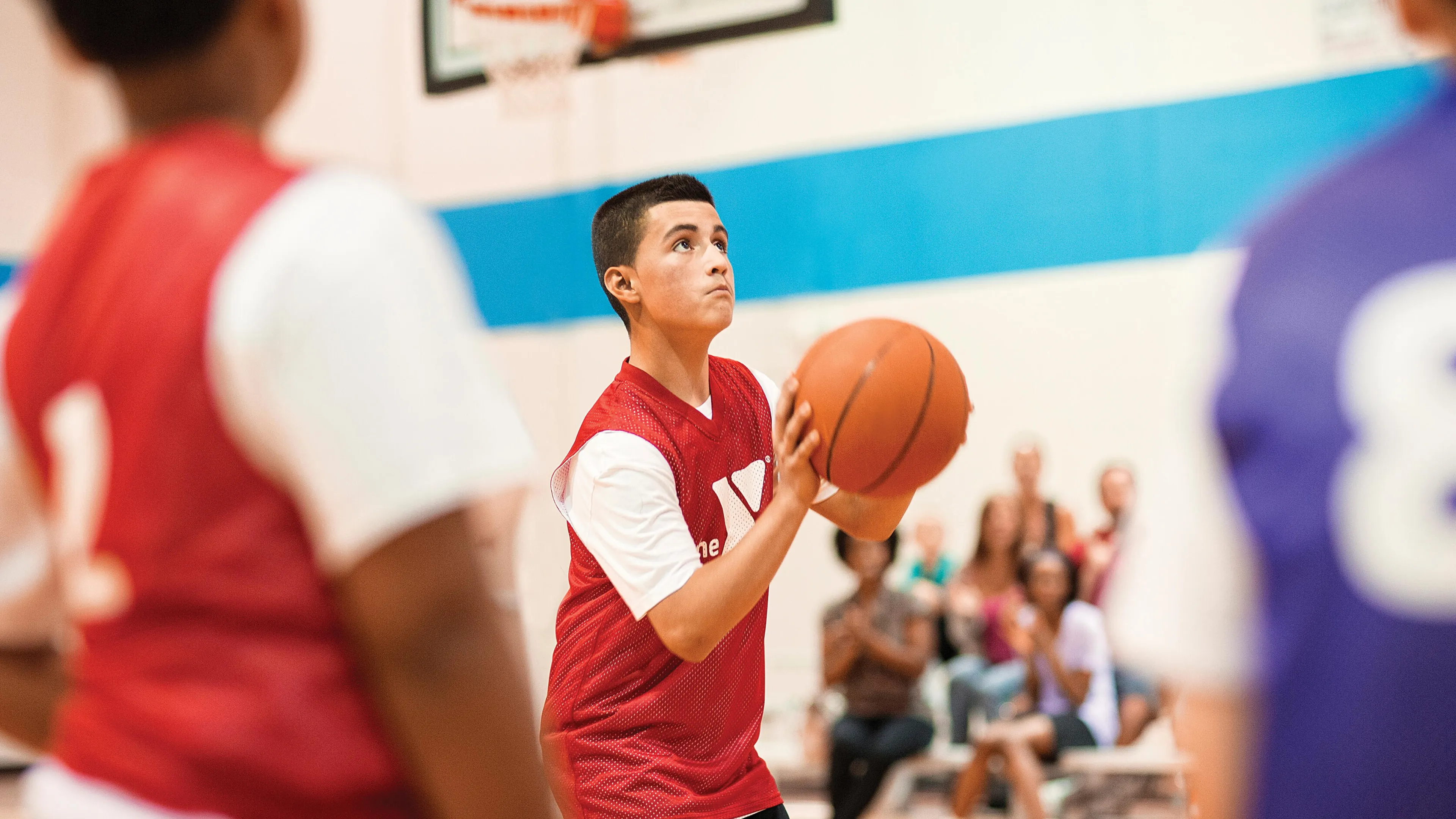 Teenager in red jersey shoots basketball in gymnasium, looking up towards the hoop. Spectators and other players blurred in background.