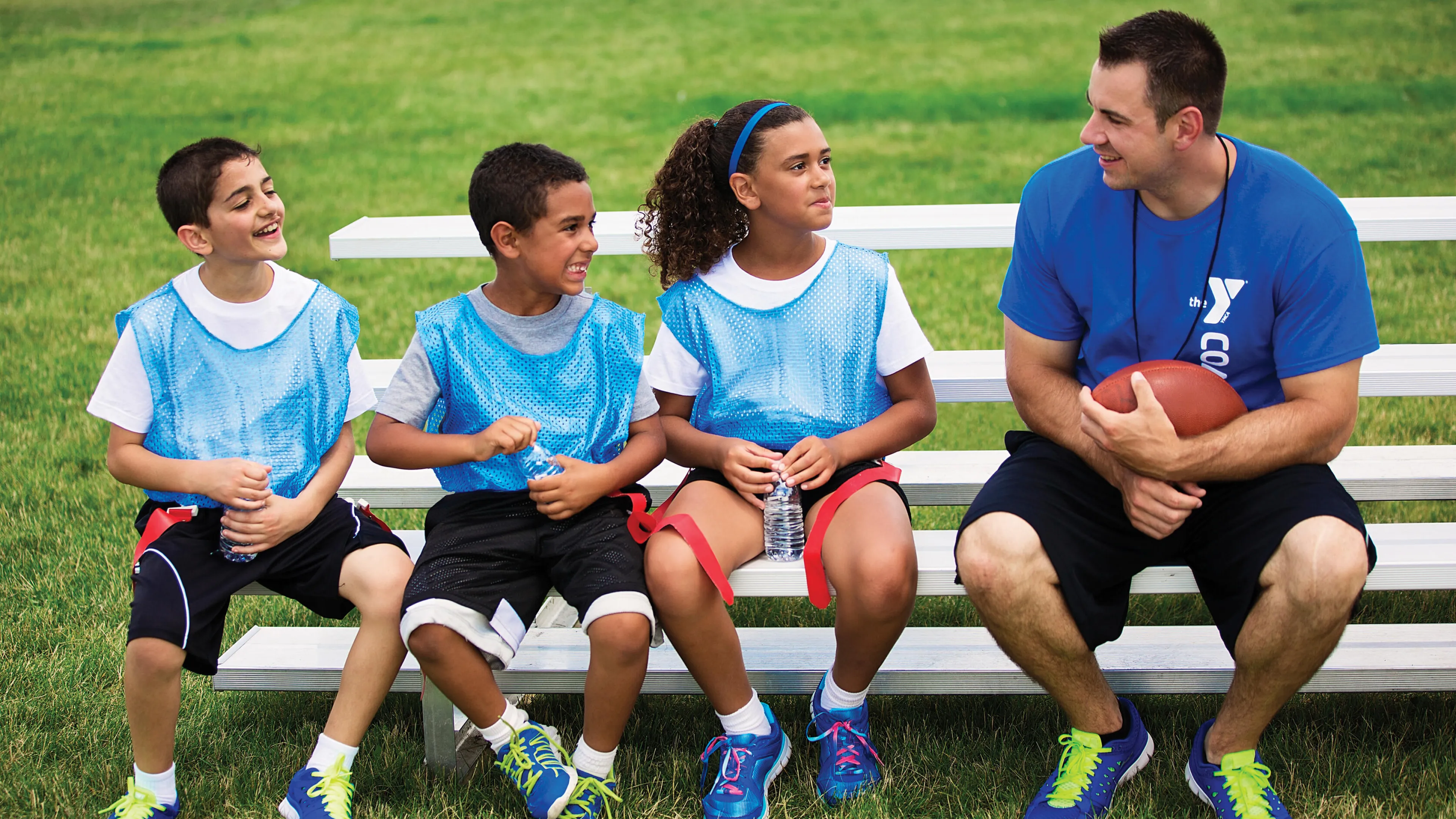 Four kids and a coach sit on a bench on a grass field. The coach holds a football. Kids wear vests and listen.