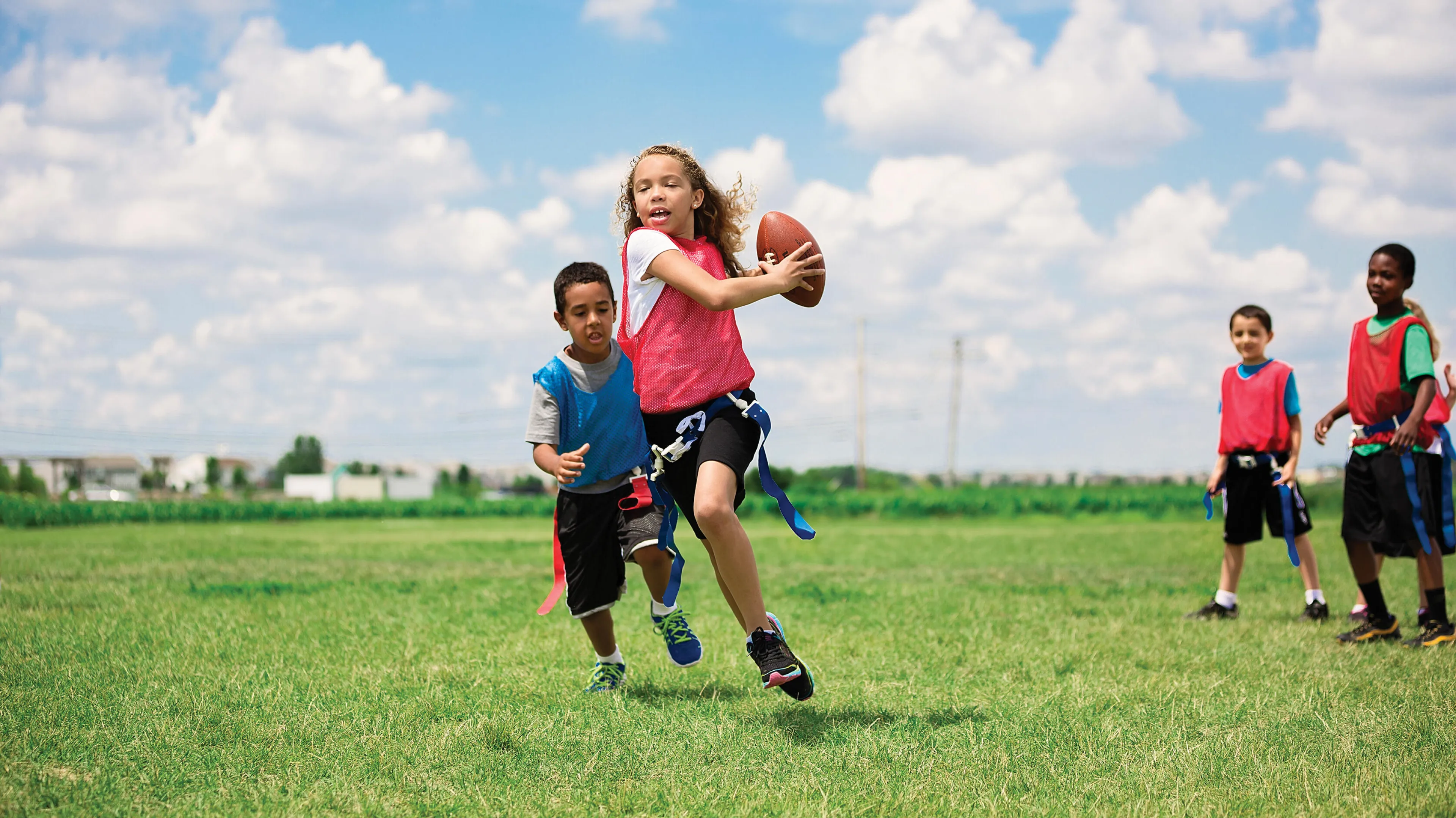 Children playing flag football on a grassy field, girl throwing a football, boy running beside her, other kids watching.