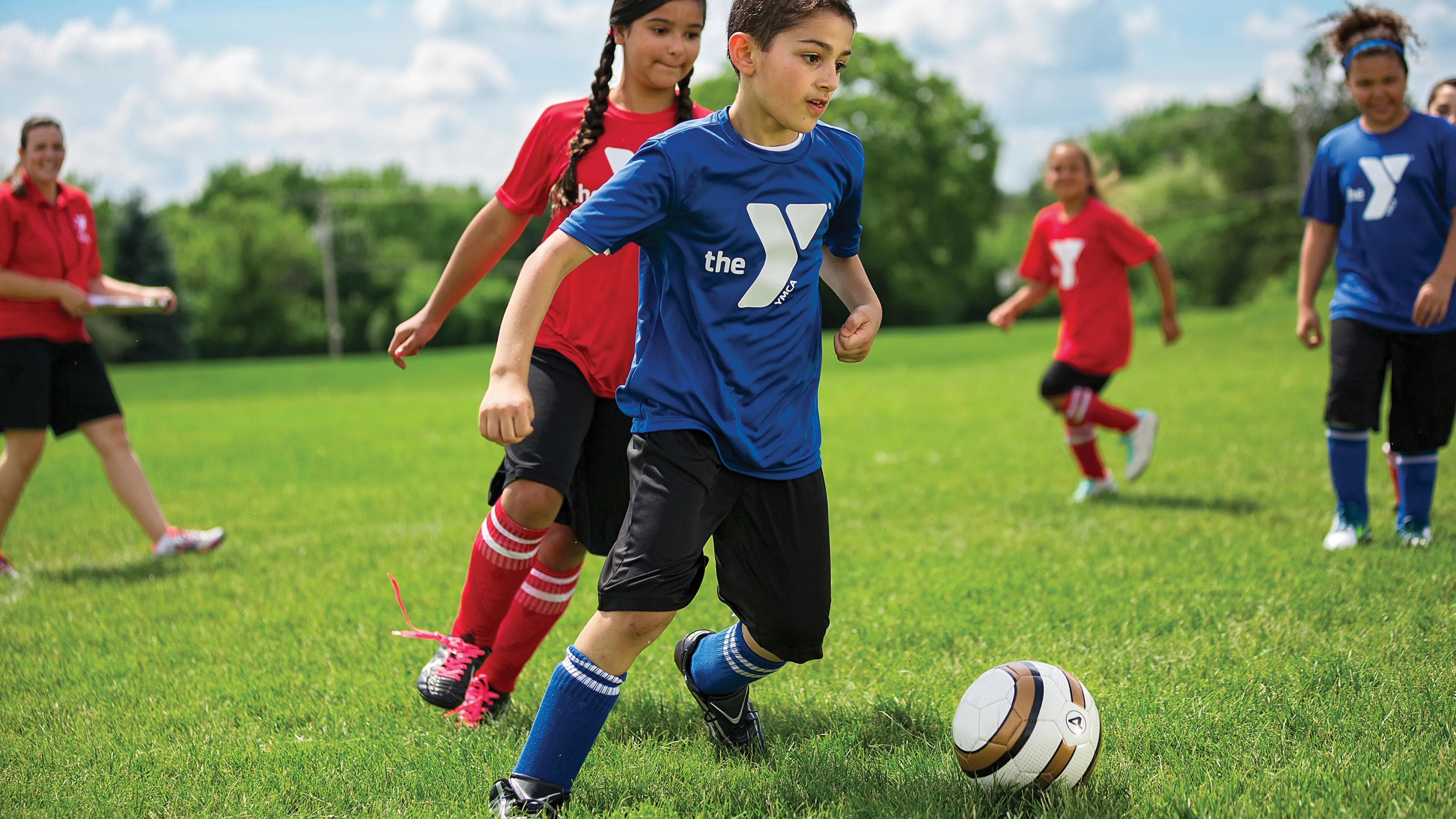 Children playing soccer on a green field; a coach observes. A boy in blue runs toward the ball while other children are playing.