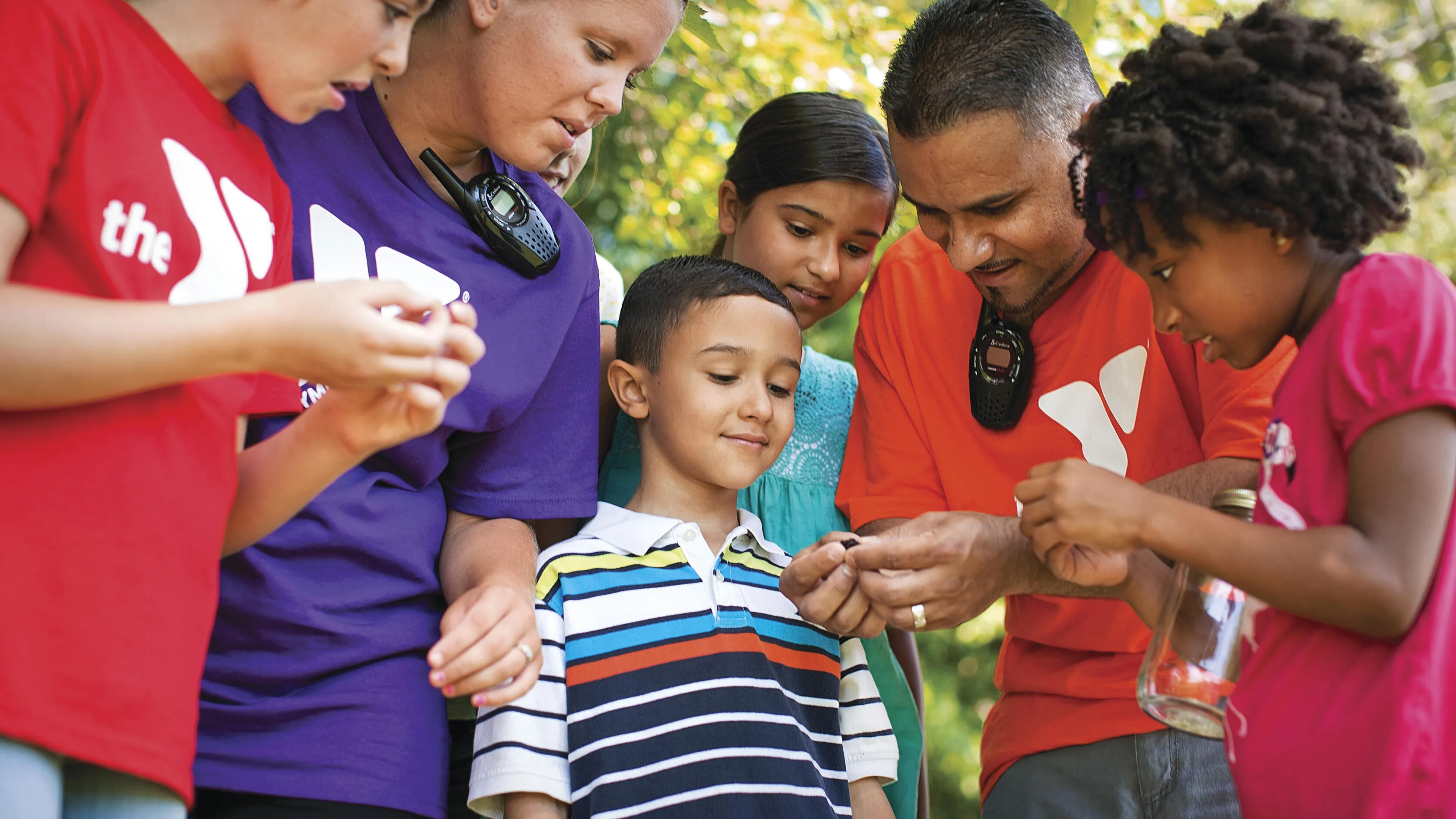 Adults and children gather around, examining something small in the open air. They are looking at an object together. The shirts have a logo.