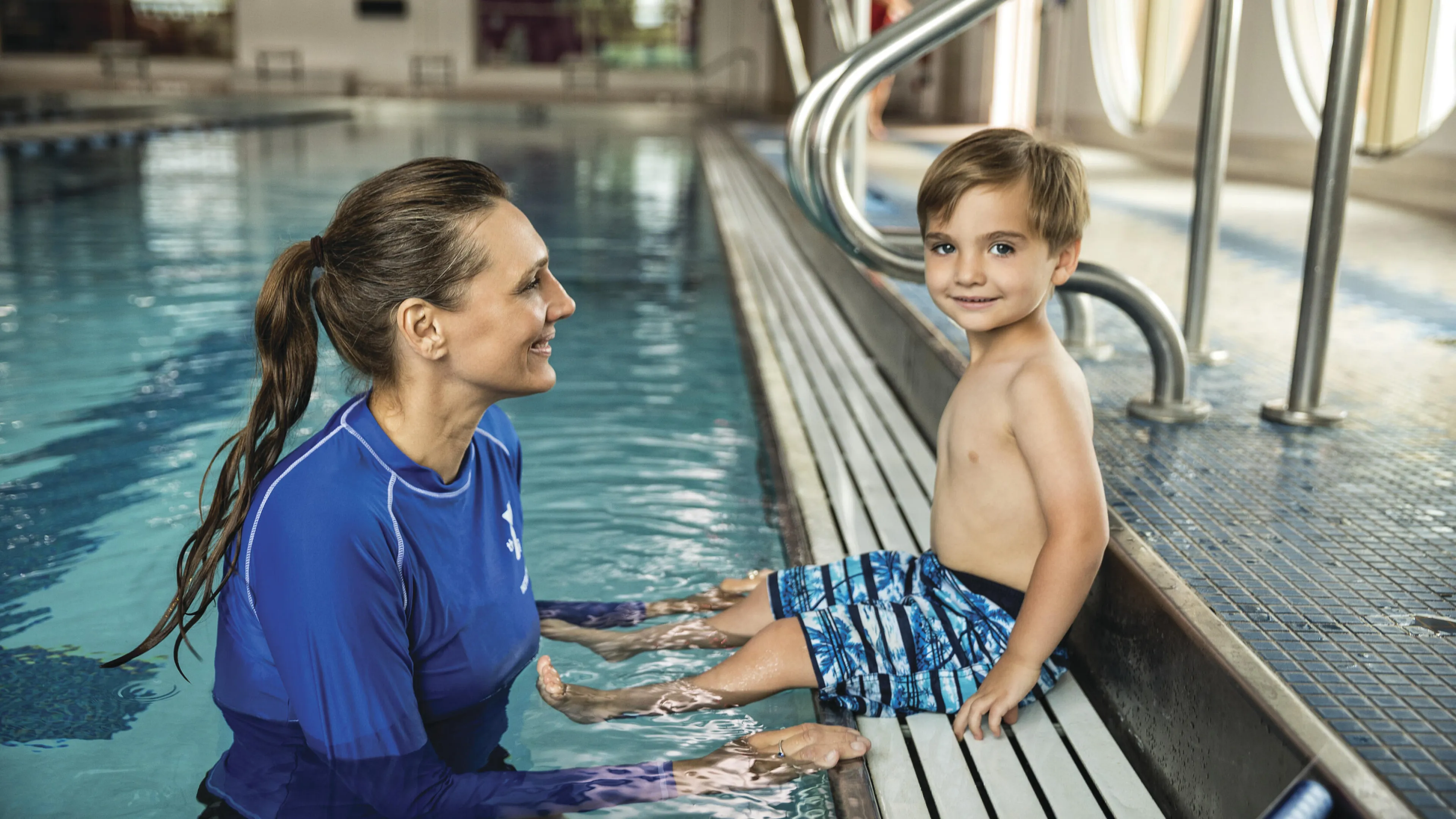 A young boy in swim trunks sits poolside, smiling at a female instructor partially submerged in the water.  They appear to be participating in a swim lesson.