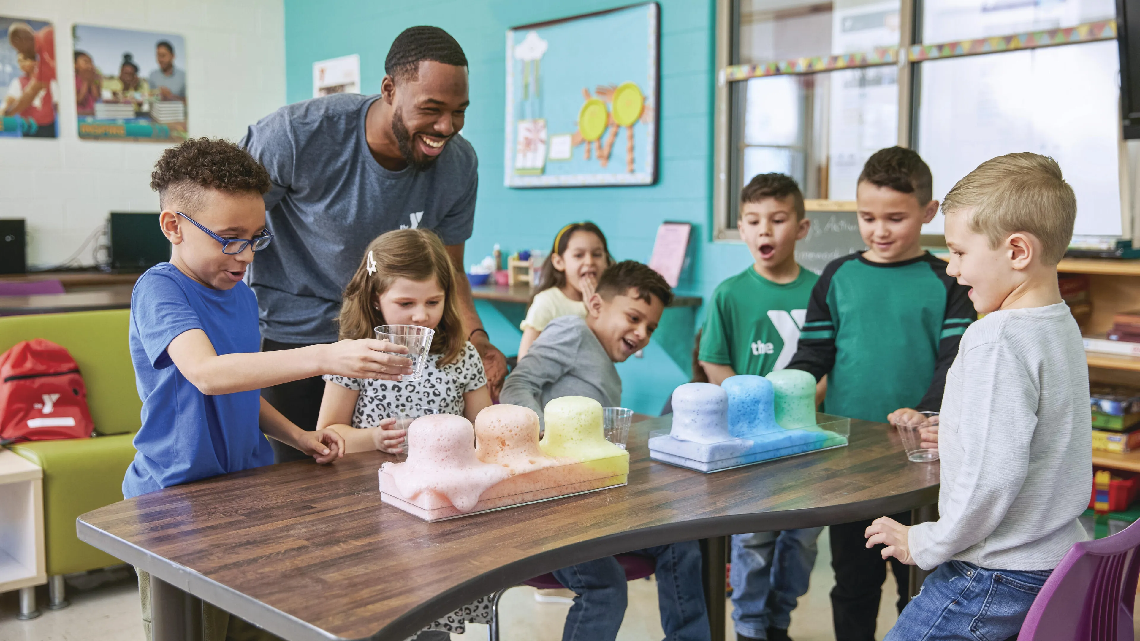 Kids conduct a science experiment with colorful substances, guided by a smiling instructor.  They're engaged and excited, working together at a table in a brightly colored classroom.