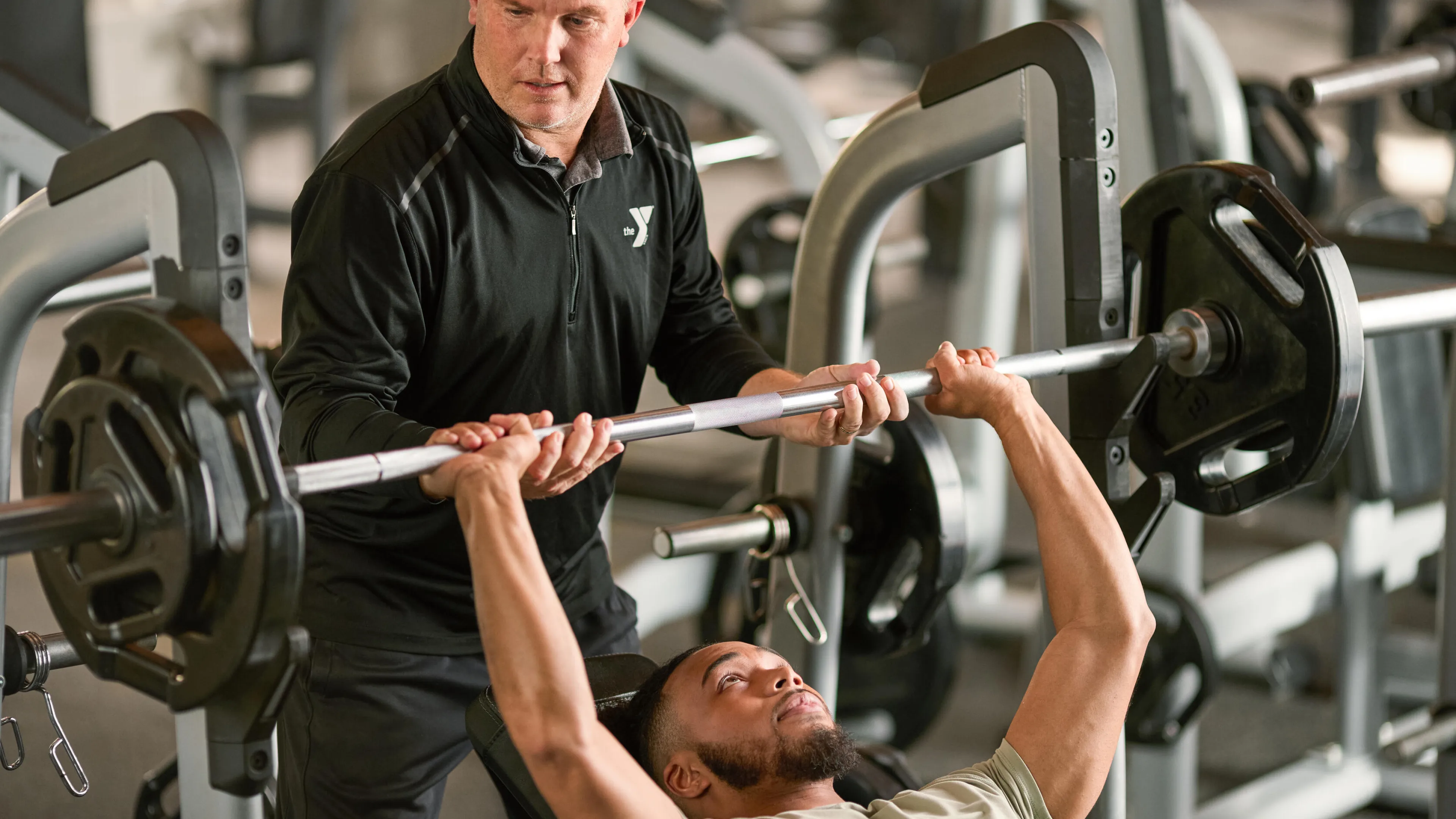 A man receives guidance from a fitness instructor while bench pressing a barbell in a gym.