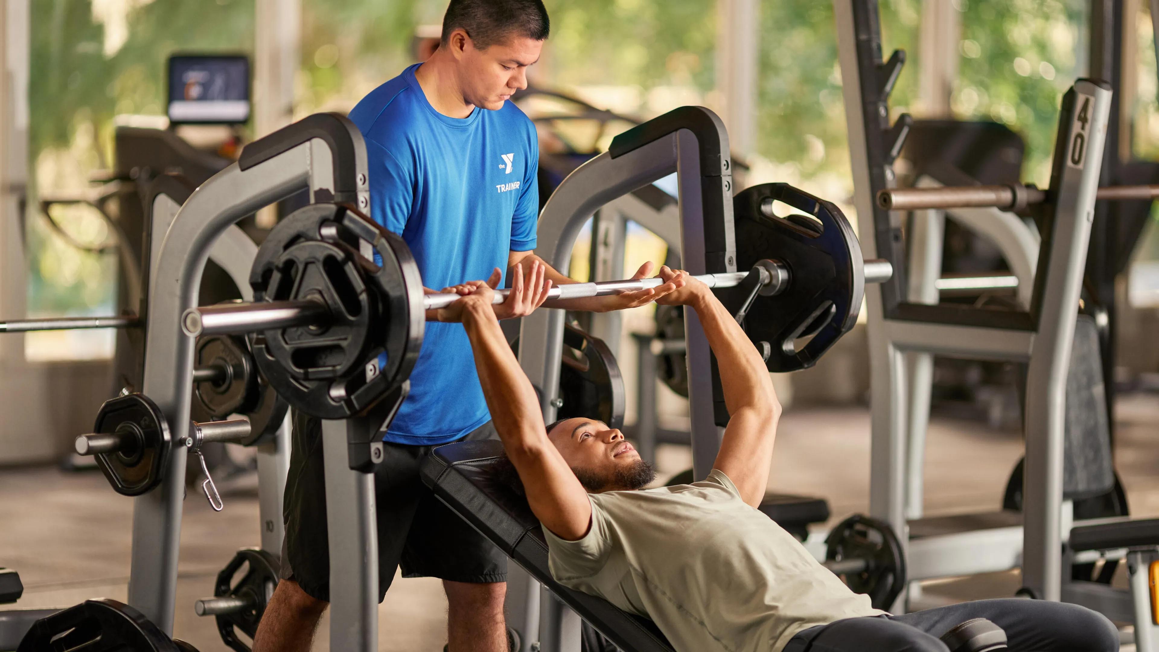A man bench presses a weighted barbell, assisted by a trainer in a bright blue shirt.  Other fitness equipment is visible in the background.