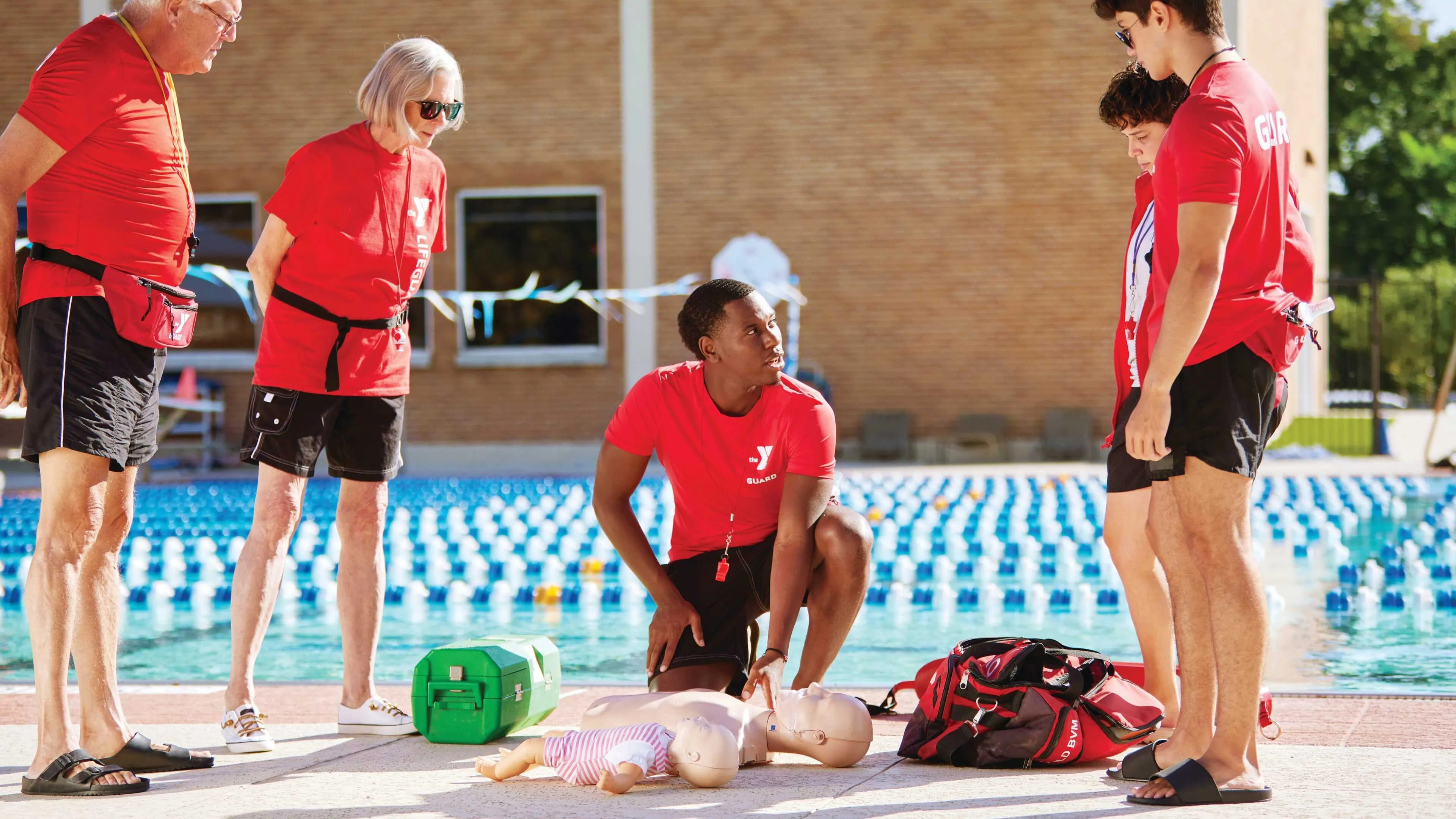 Lifeguards in red shirts practice CPR on a training dummy by the pool with others watching. A green first aid kit and a red bag lay nearby.