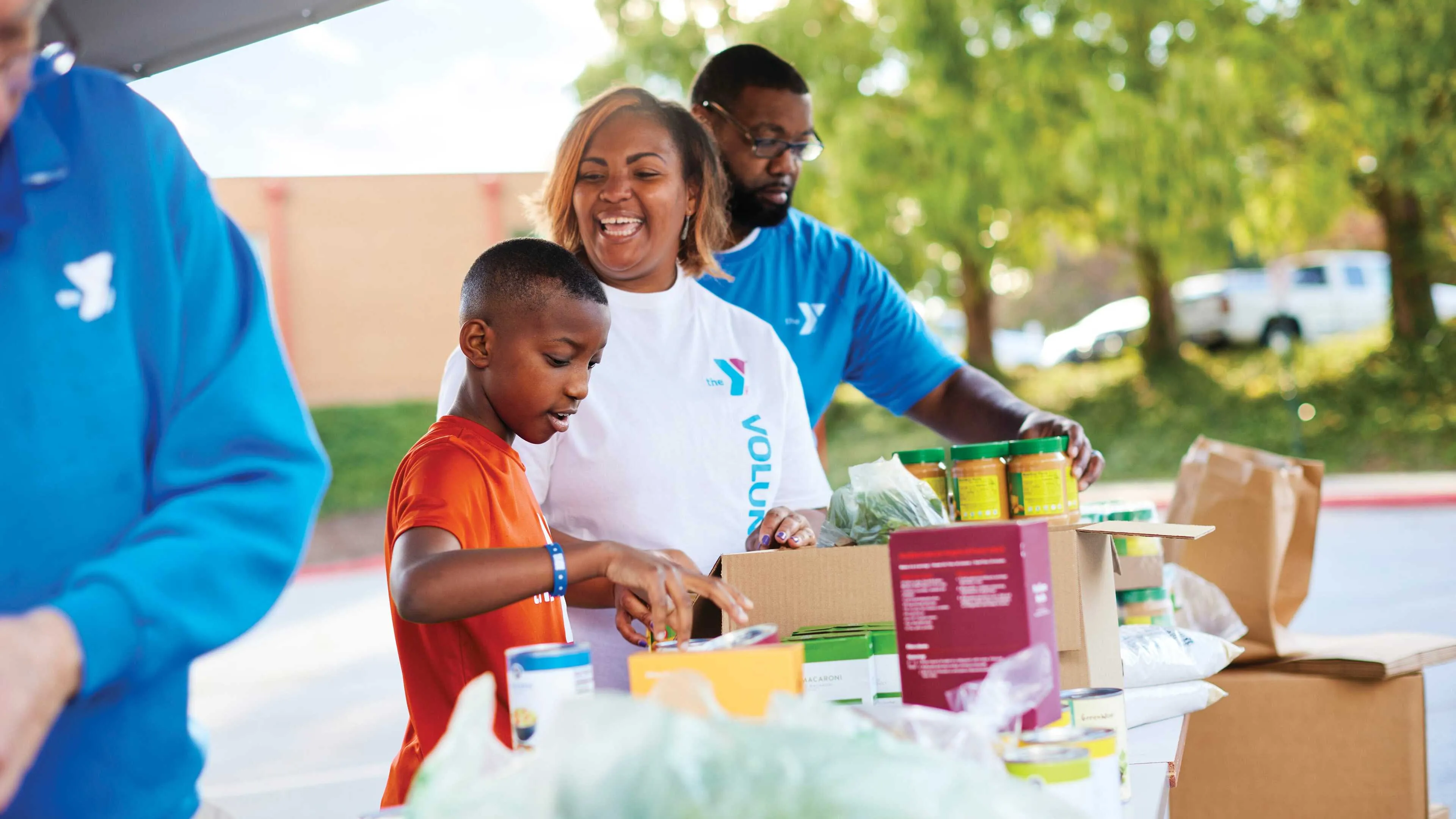 Volunteers and a young boy packing food in boxes at an outdoor community event; they are sorting items and smiling.